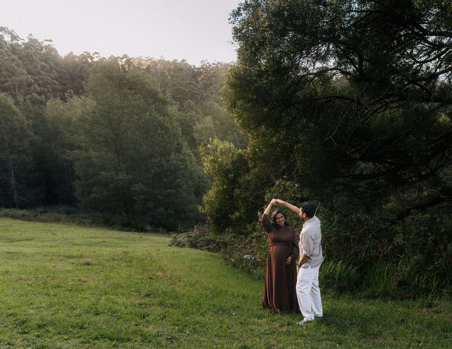 Couple dancing hand in hand on a lush green meadow, woman in a rust dress and man in white, with tall forest trees behind them.