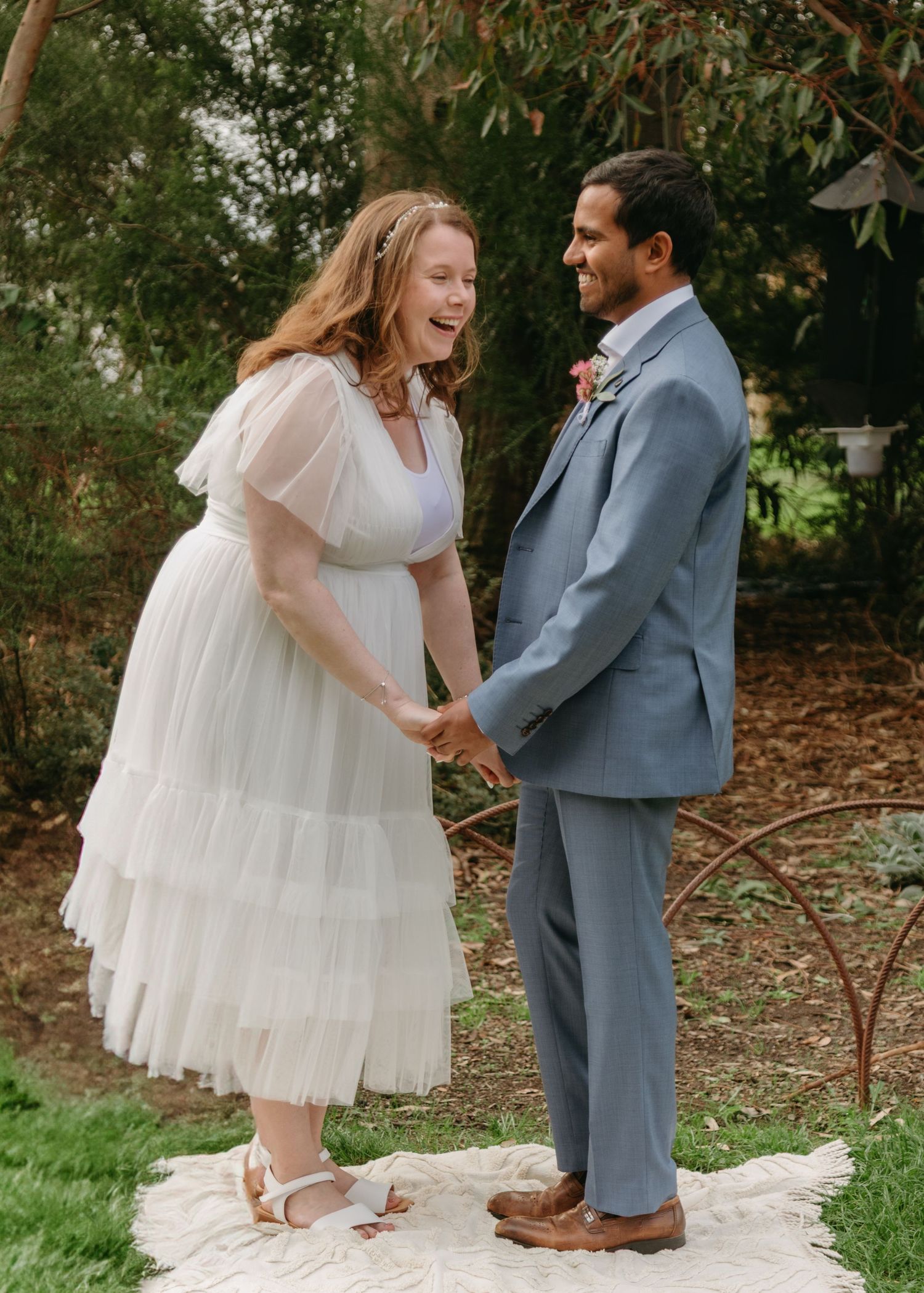 Bride in white tiered tulle dress and groom in blue suit laughing joyfully while holding hands.