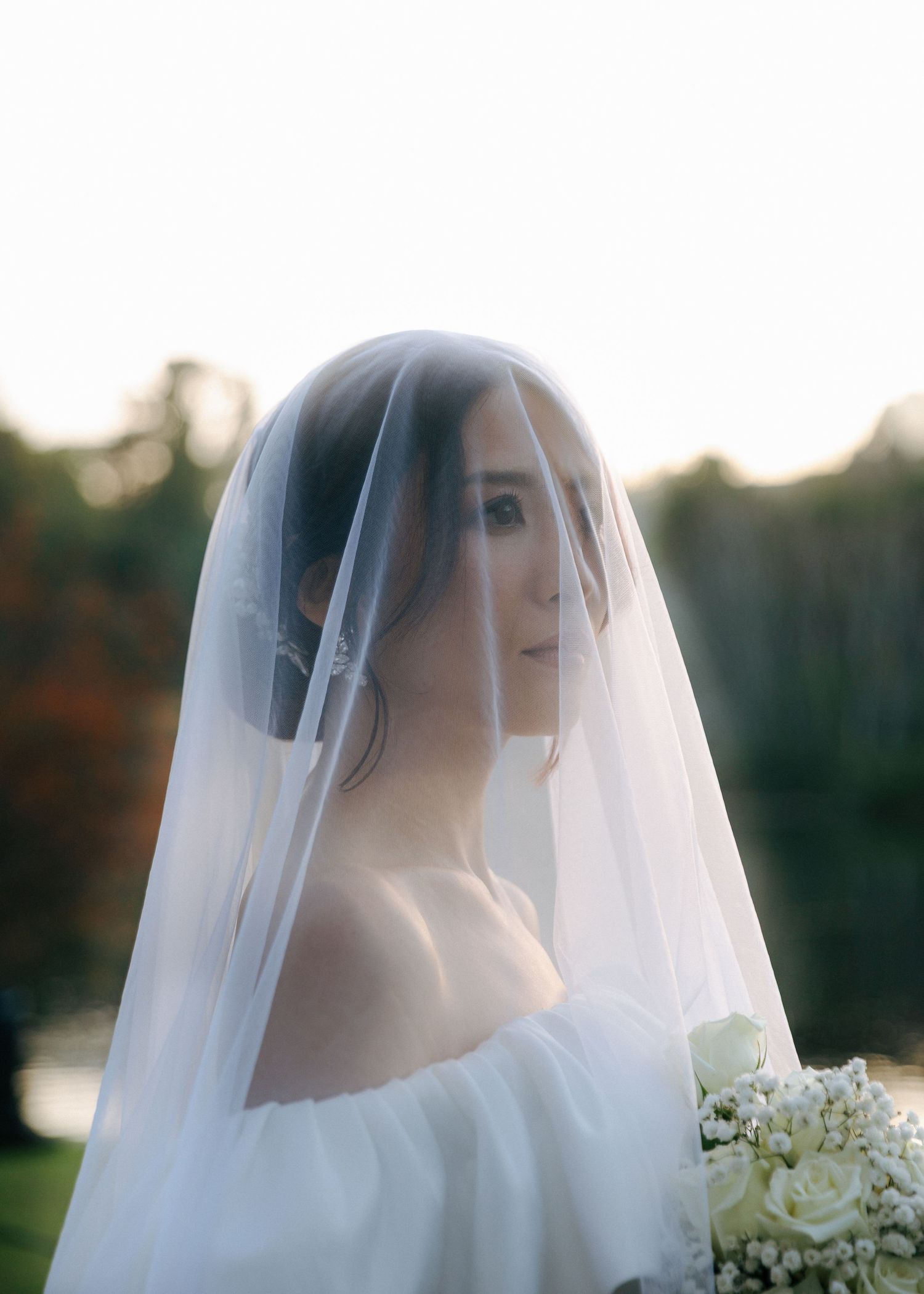 Bride in an off-shoulder wedding dress and veil holds a white bouquet, bathed in soft backlit sunlight.