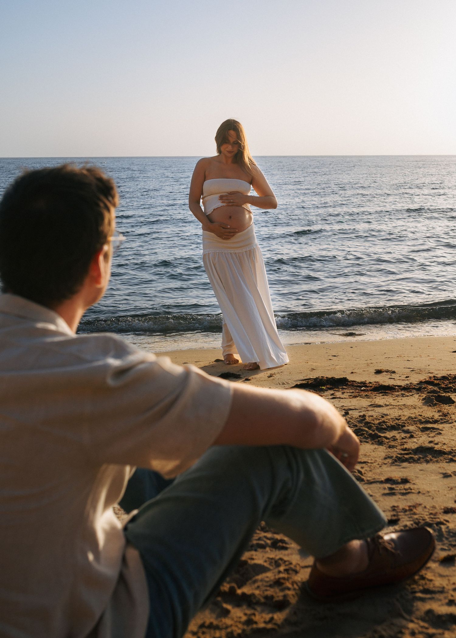 Pregnant woman in white dress holds belly on beach while man sits on sand watching her at sunset.