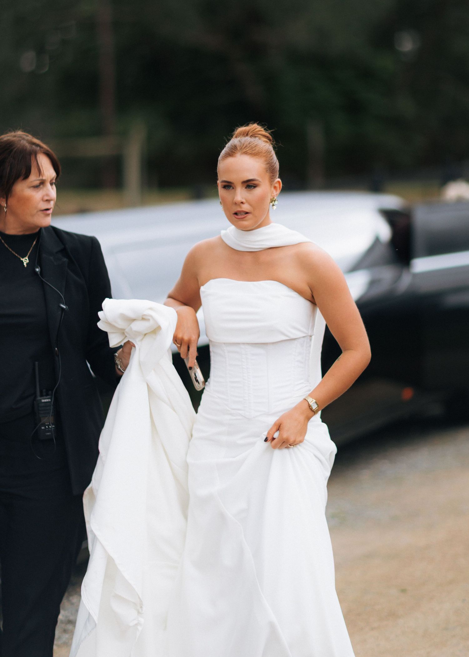 Bride in strapless white corset gown with scarf walks beside assistant near limousine.