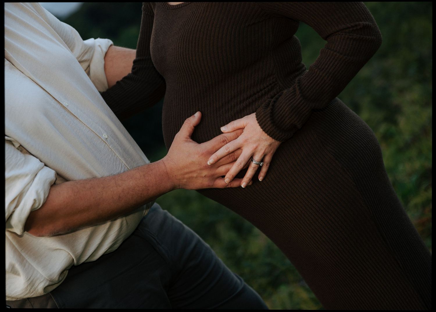 Pregnant woman in brown outfit with partner's hands cradling baby bump.