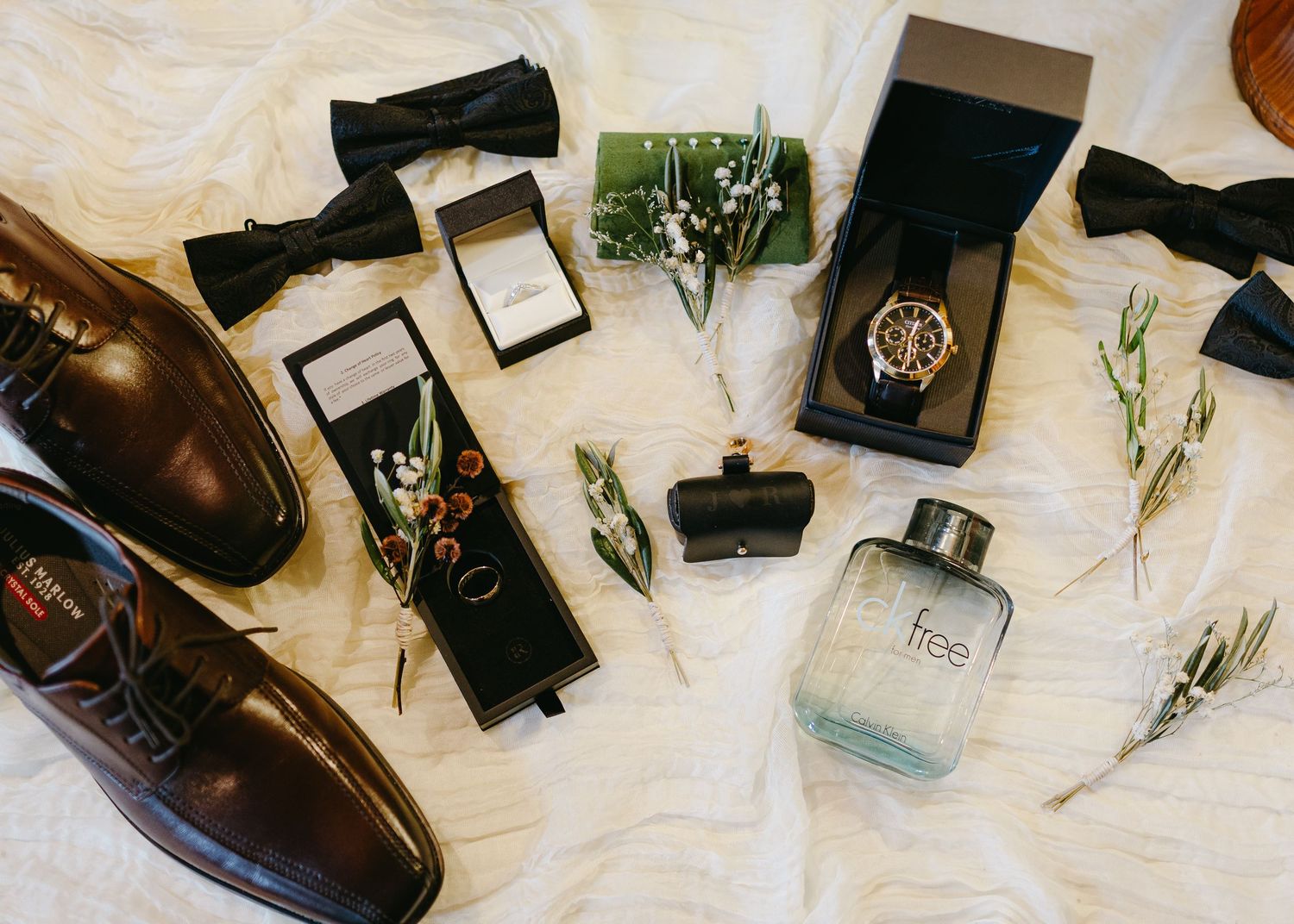 Groom wedding flat lay with bow tie, watch, rings, cologne, and brown leather shoes on white fur.
