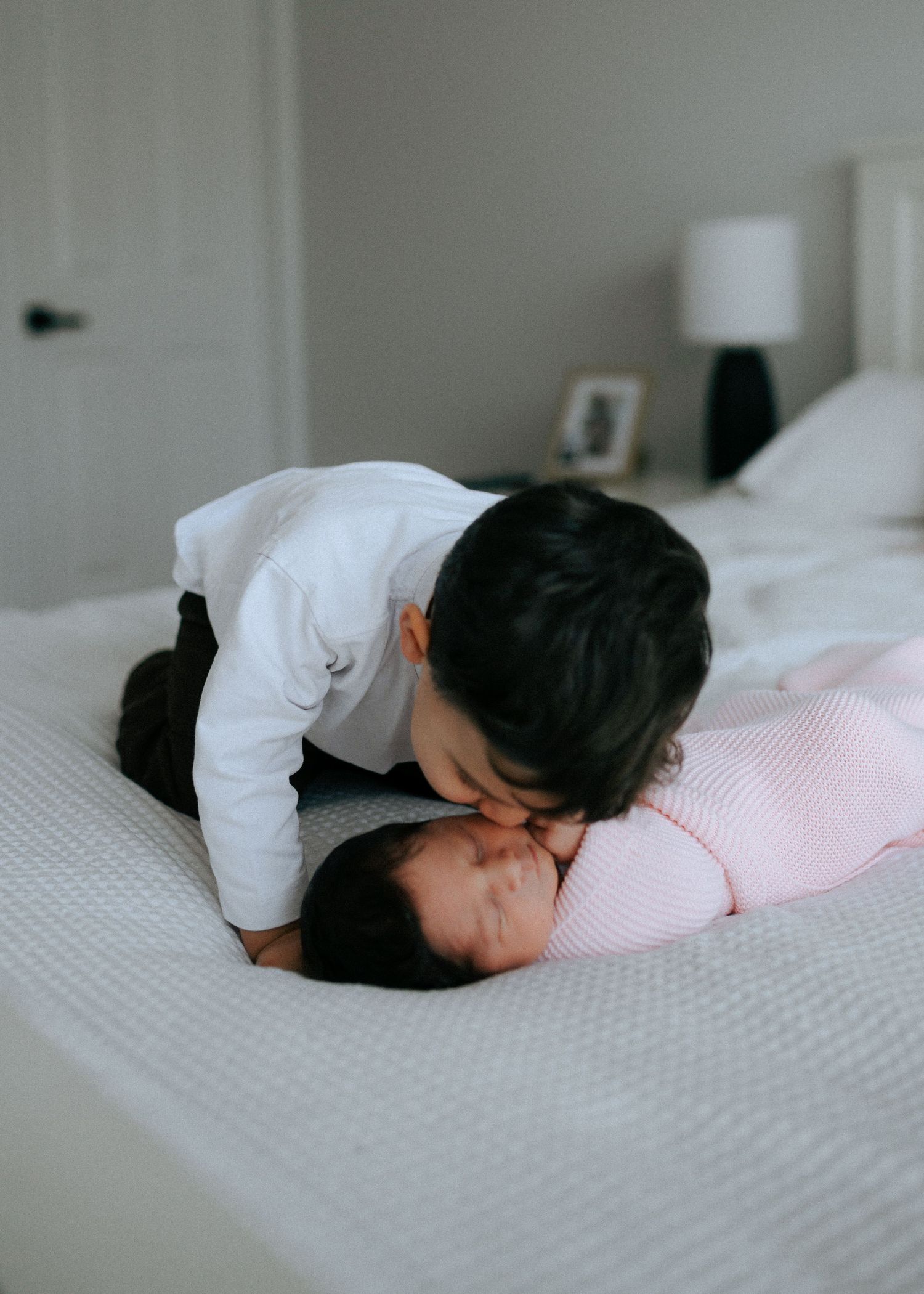 Young toddler boy in white shirt gently kissing newborn baby sibling on white bed.