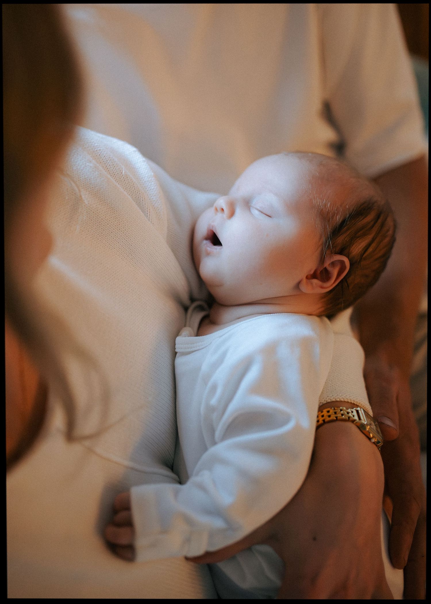 Sleeping newborn in white onesie cradled in parent's arms with gold watch visible.