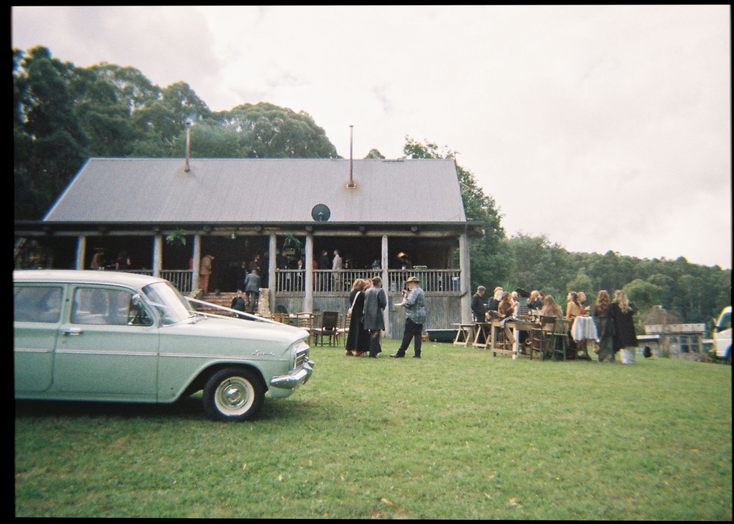 Wedding guests gather outside a rustic venue building beside a vintage light blue classic car on grass.