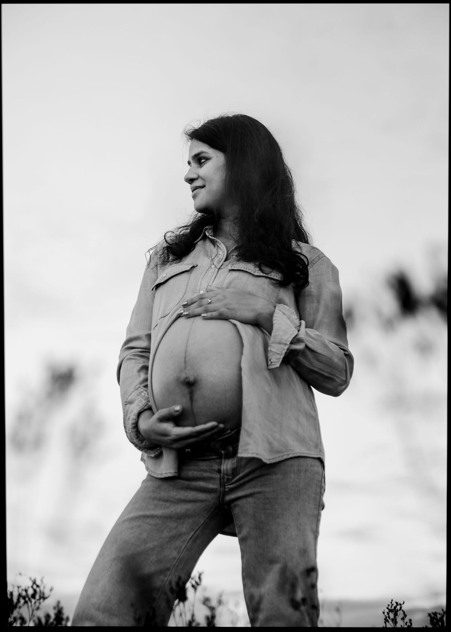 Black and white portrait of pregnant woman in denim shirt cradling baby bump outdoors.