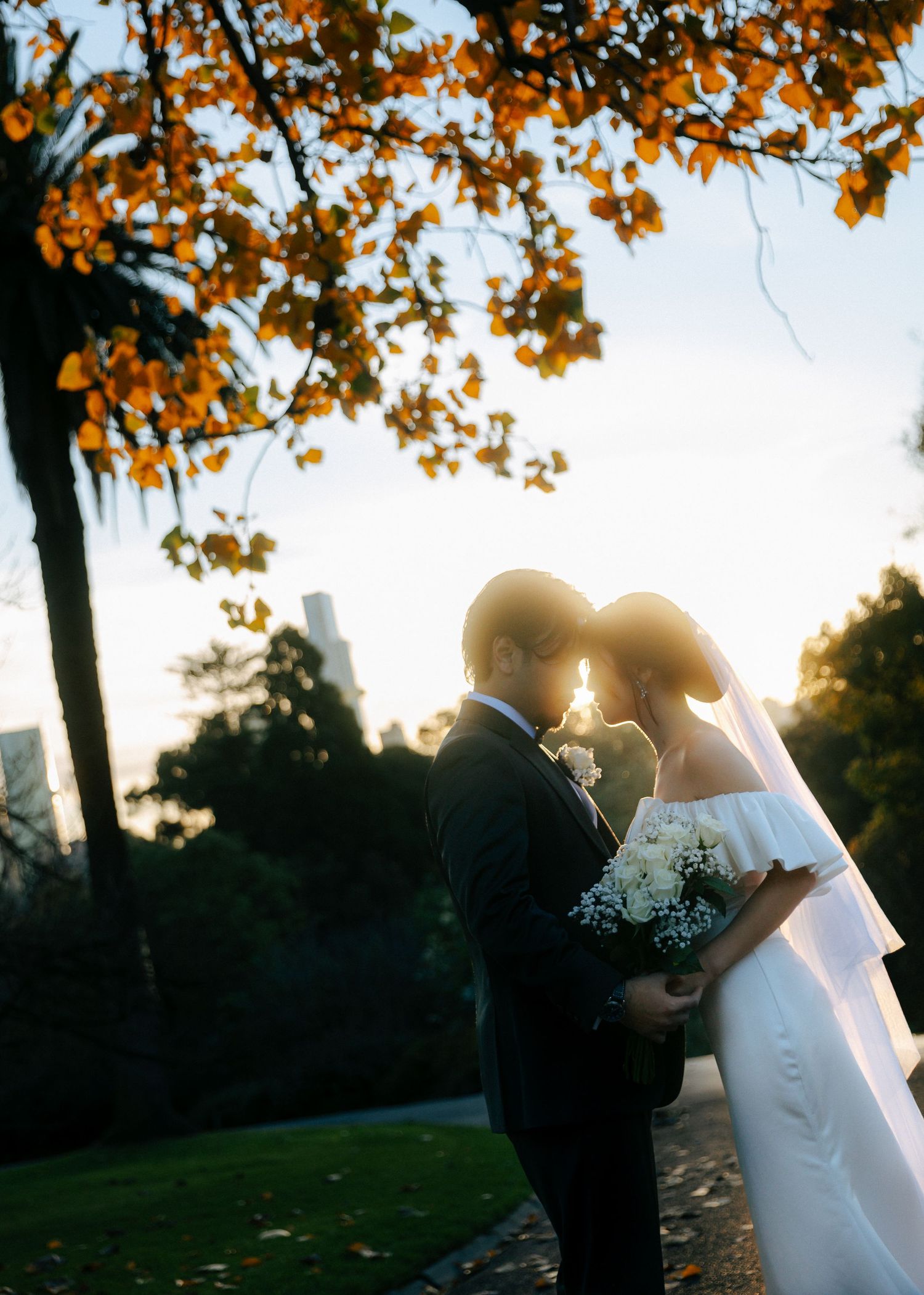 Bride and groom sharing a romantic moment backlit by golden sunset light beneath autumn foliage in a park.