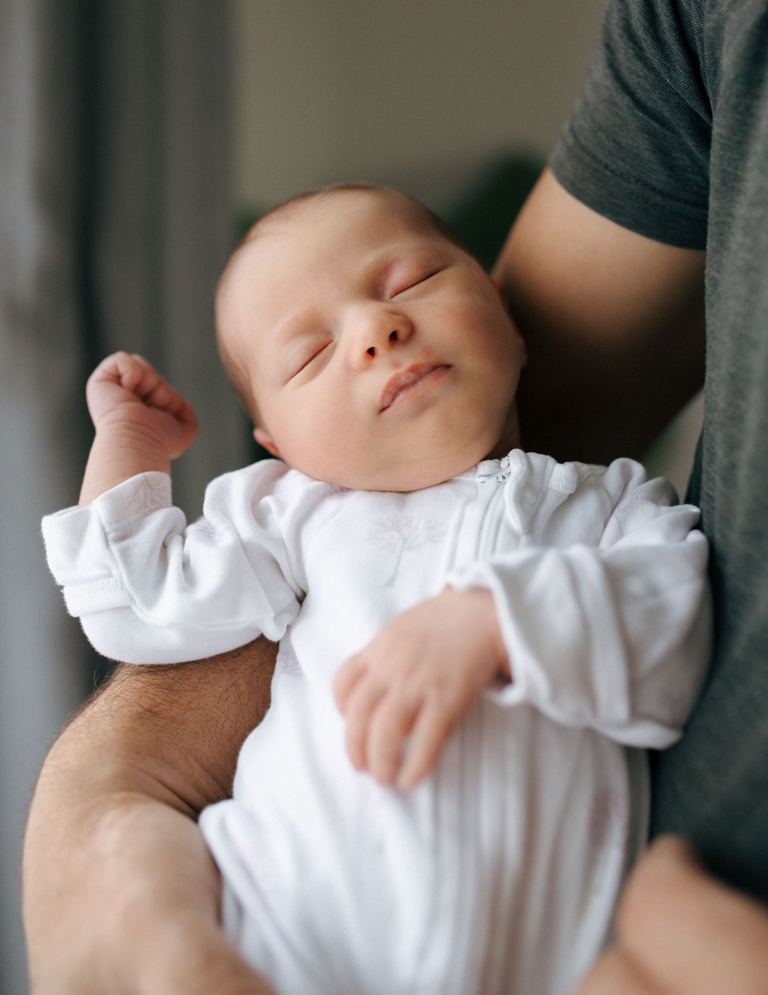 Newborn baby sleeping peacefully in white onesie cradled in fathers arms.