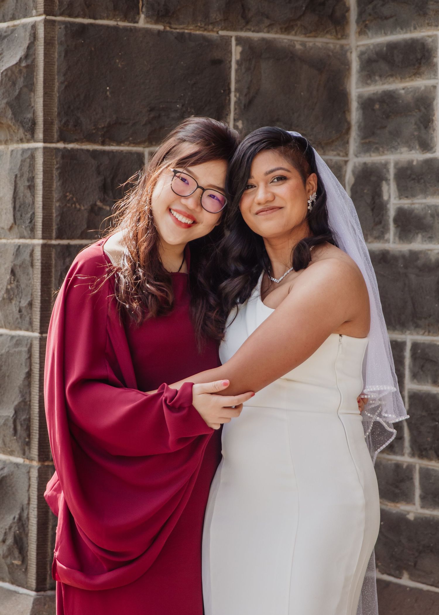 Bride in white gown and veil poses with bridesmaid in burgundy dress against stone wall.