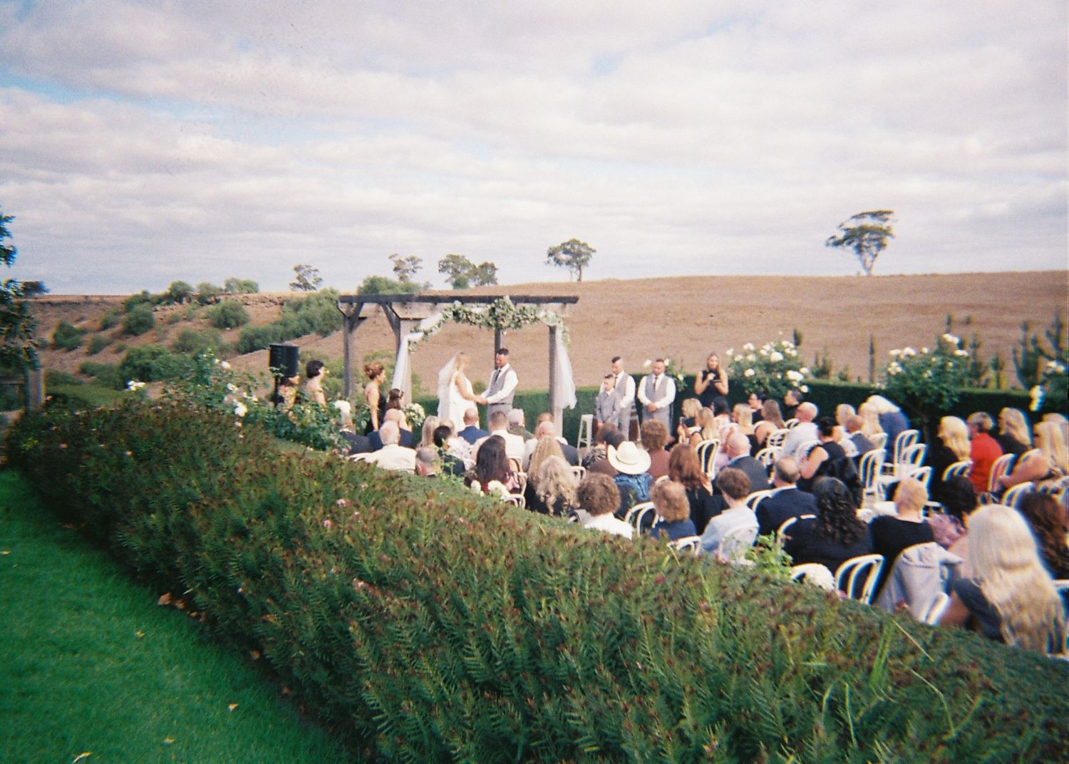 Outdoor wedding ceremony with guests seated along a manicured hedge with open farmland in the background.