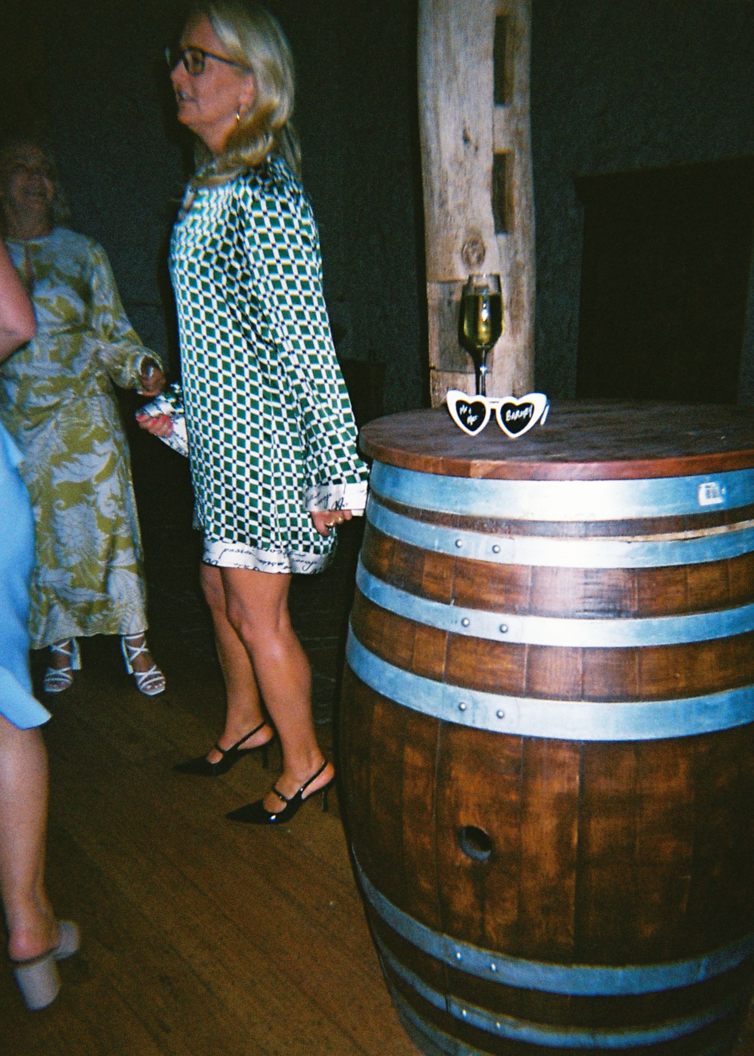 Woman in green patterned dress standing beside a wooden wine barrel at a wedding reception.