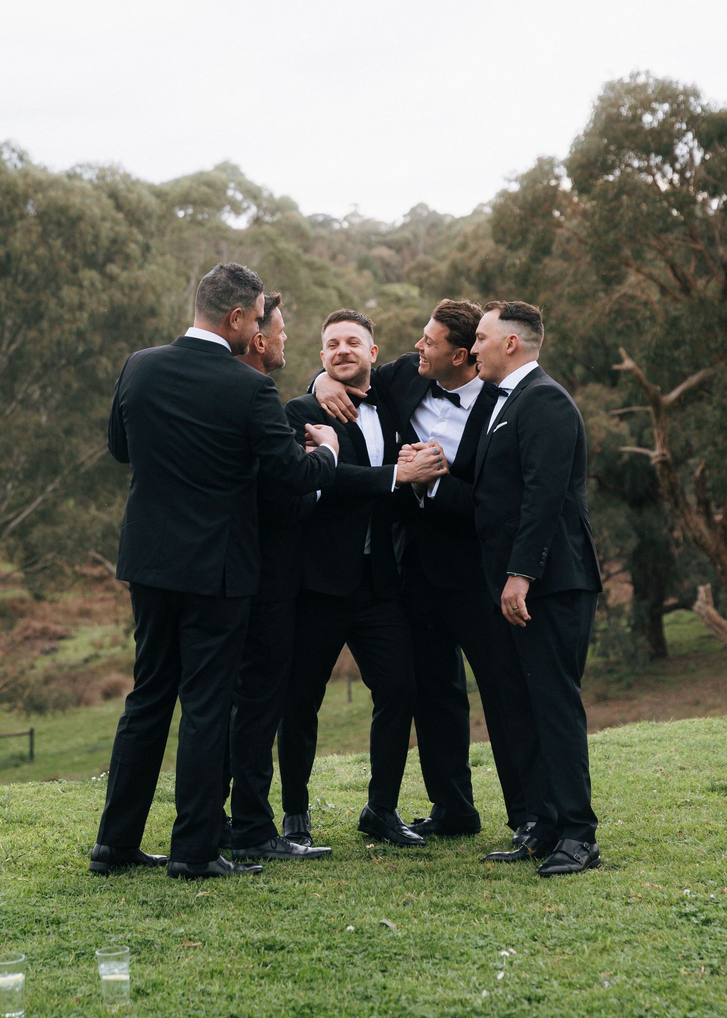 Groom and groomsmen in black tuxedos sharing a joyful embrace outdoors on a green lawn.
