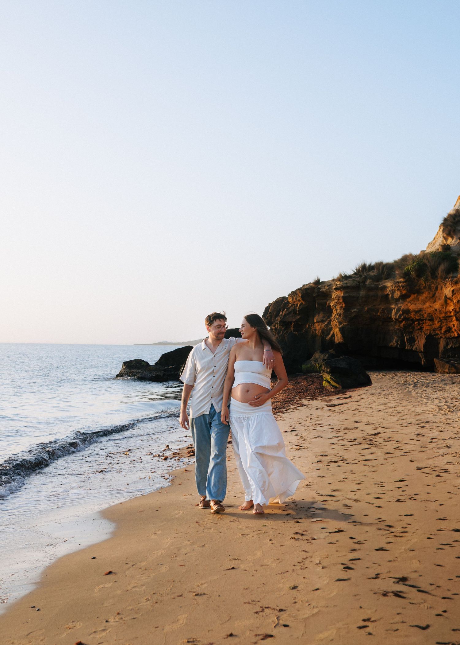 Couple walking hand-in-hand on a sandy beach at golden hour with rocky cliffs in the background.