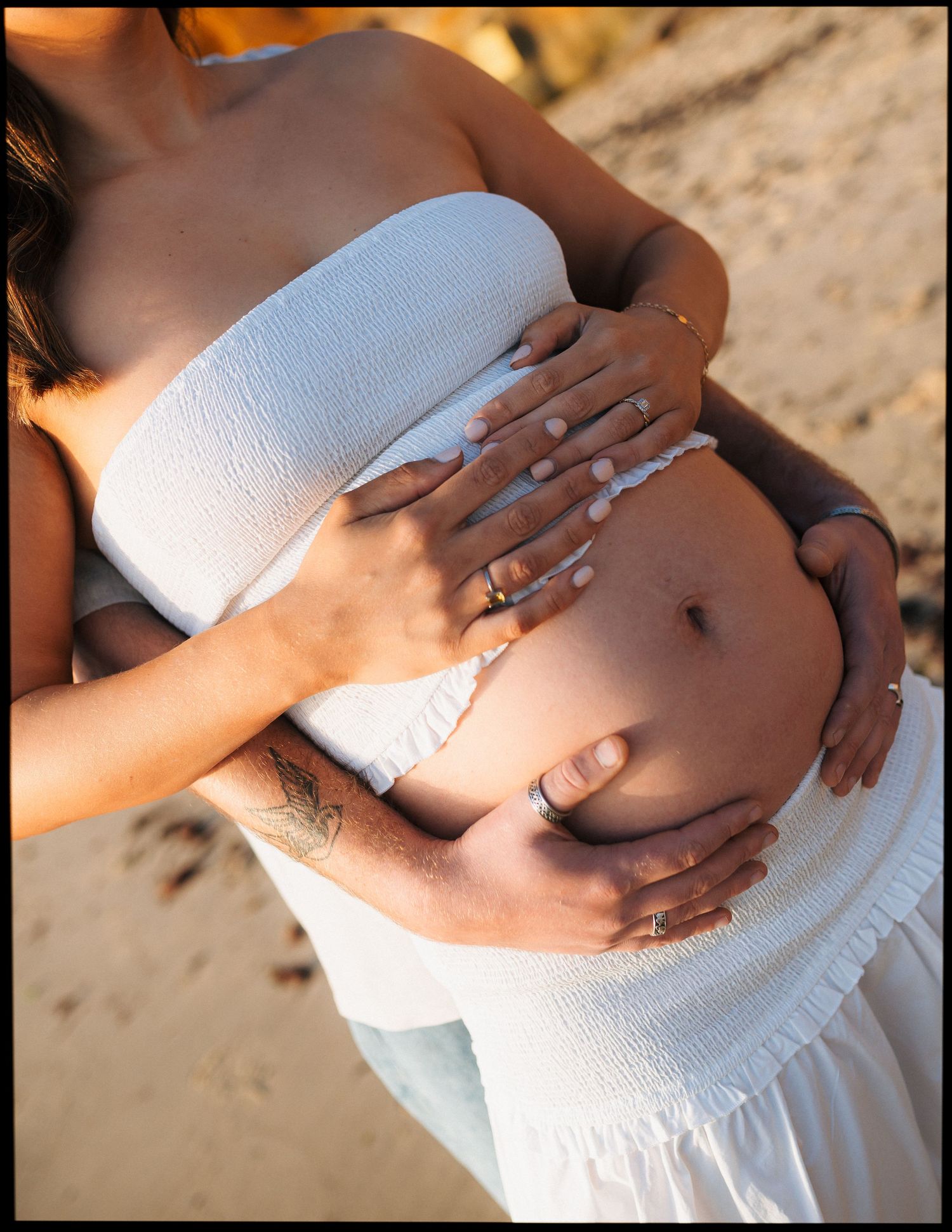 Pregnant woman in white strapless dress with partner's hands cradling her bare baby bump on a sunny beach.