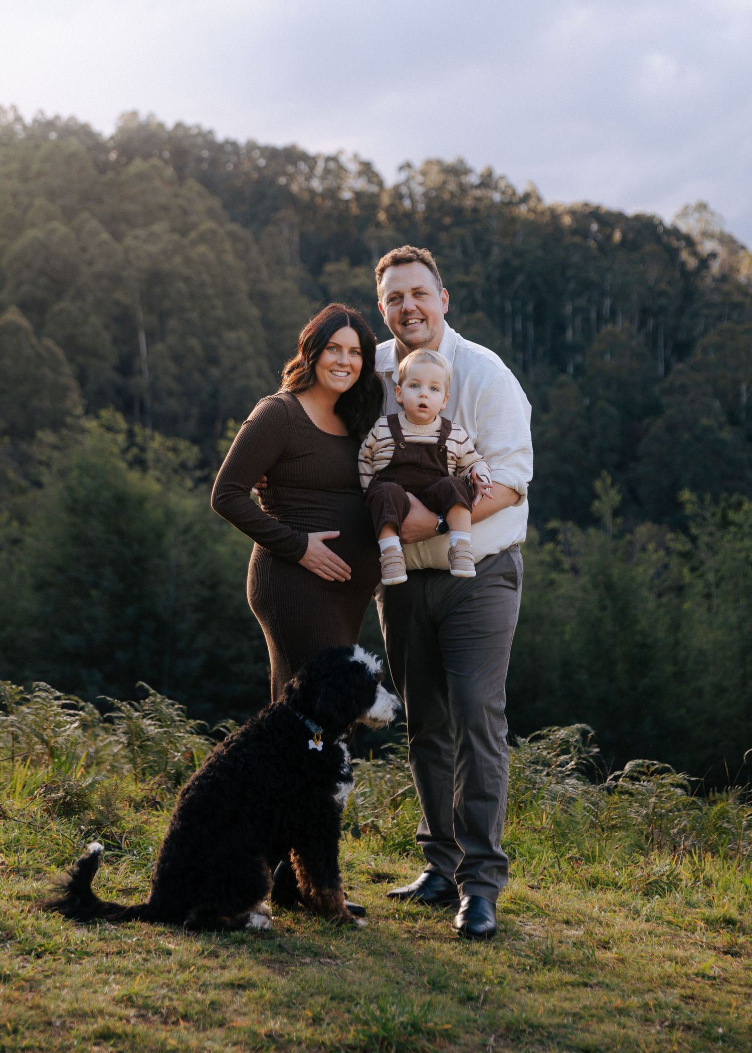 Family portrait with pregnant mother, father holding toddler, and black and white dog on green hillside.