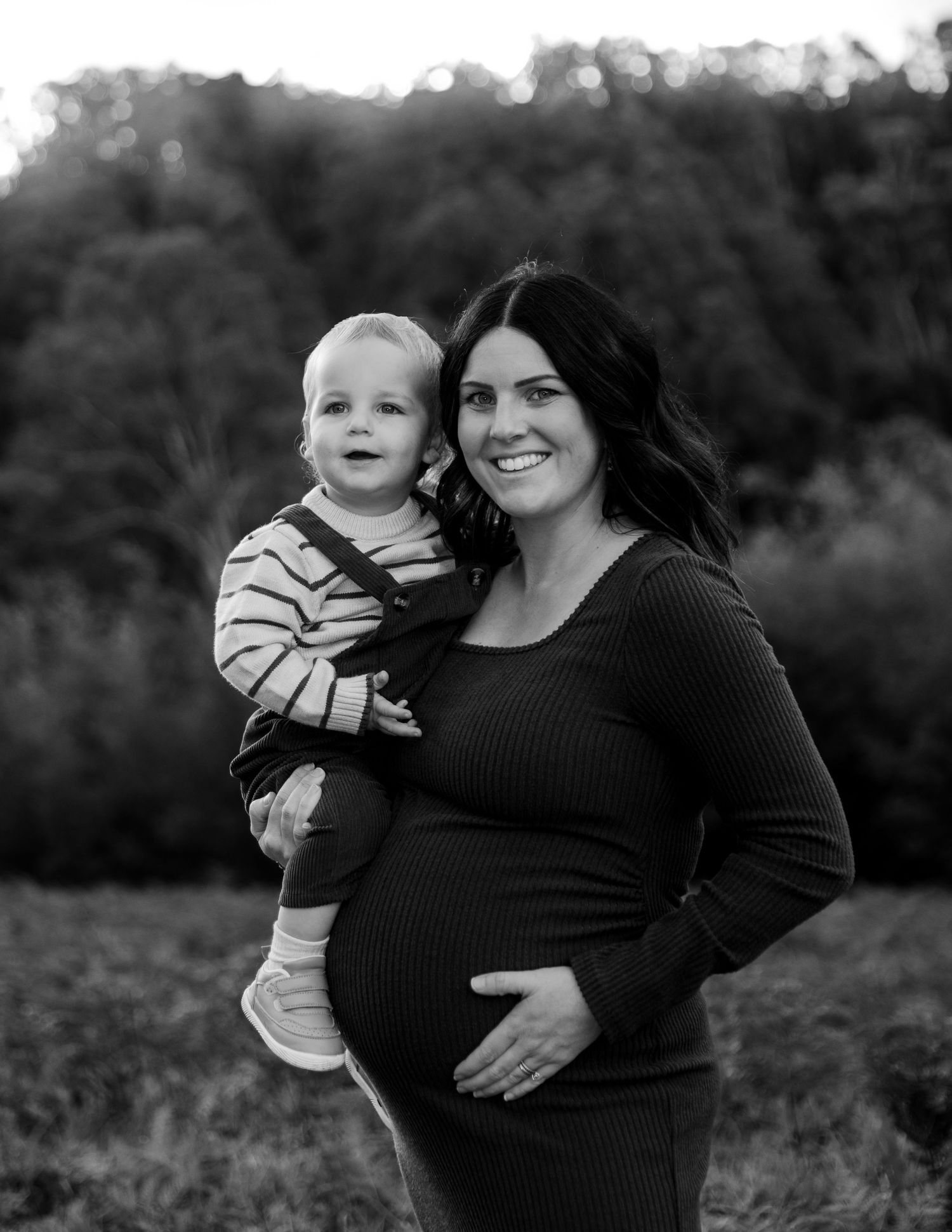 Black and white maternity photo of woman holding smiling toddler outdoors in a field.