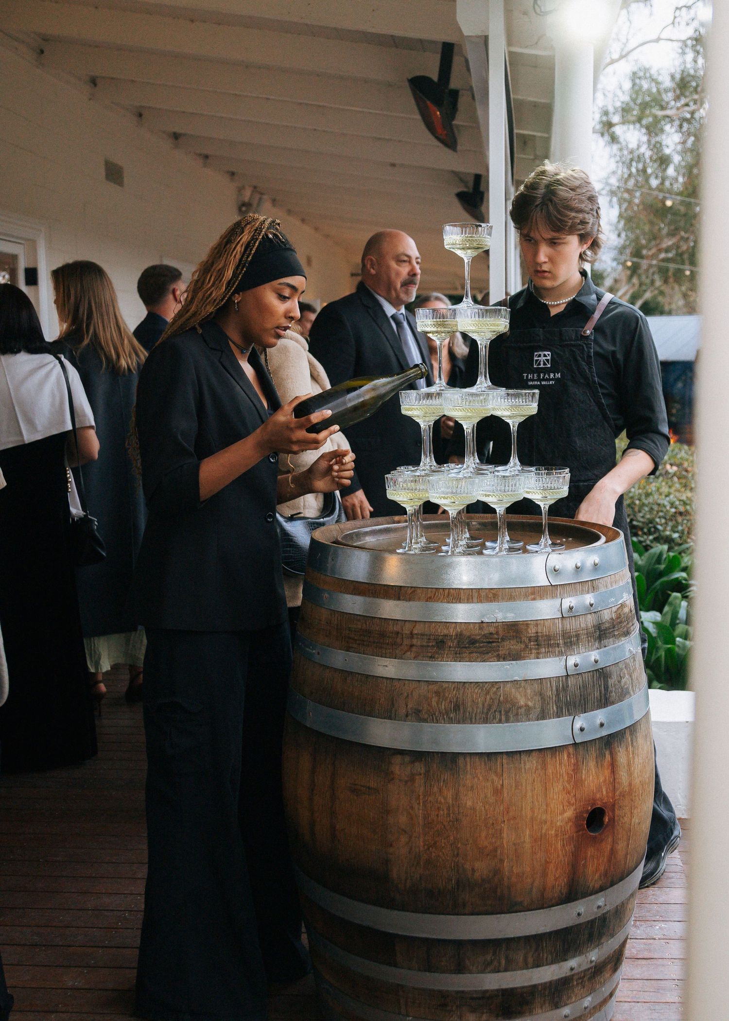 Staff pouring champagne tower on wooden wine barrel at elegant winery event.