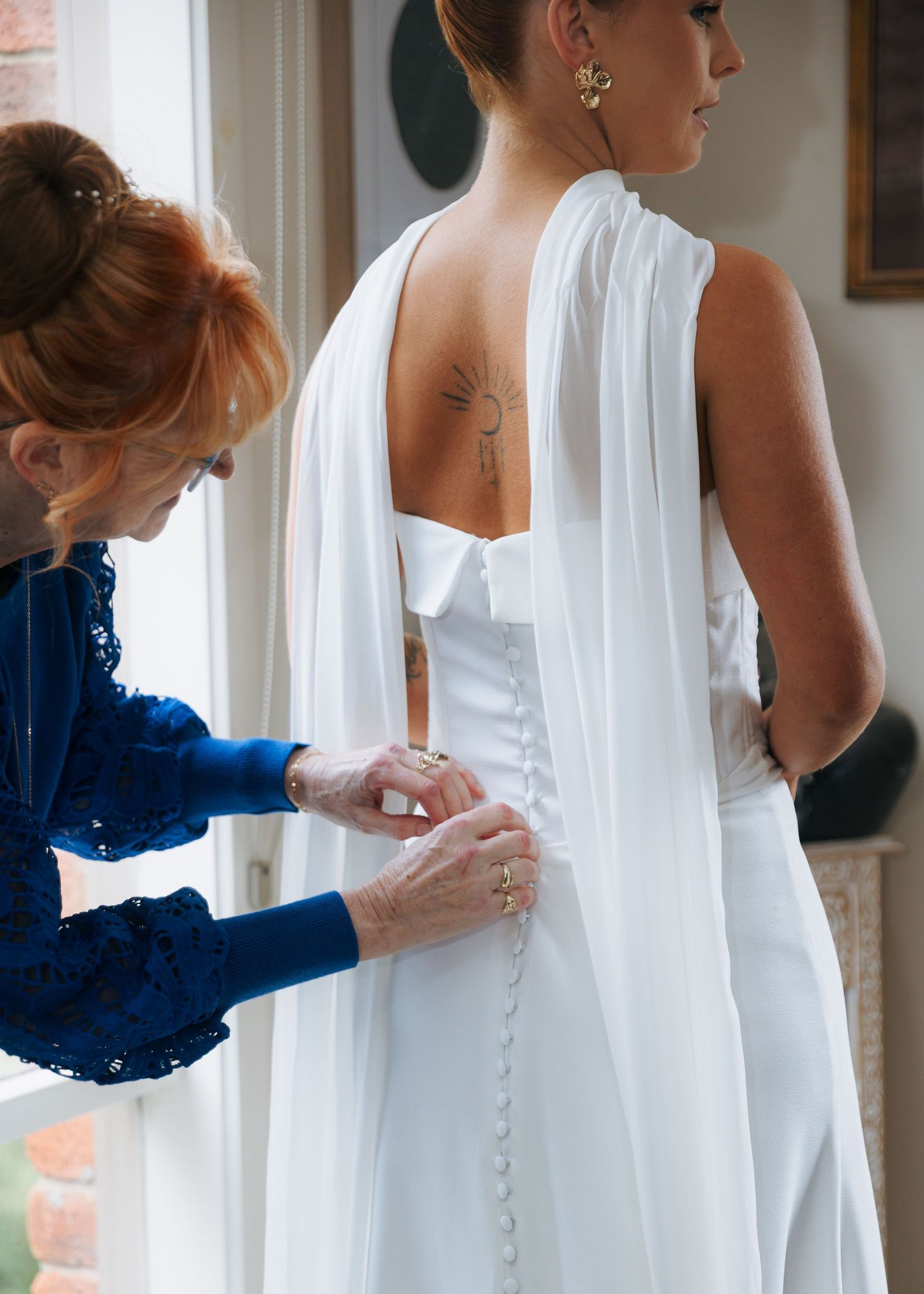 Mother fastening row of buttons on back of daughters elegant white wedding gown with flowing cape.