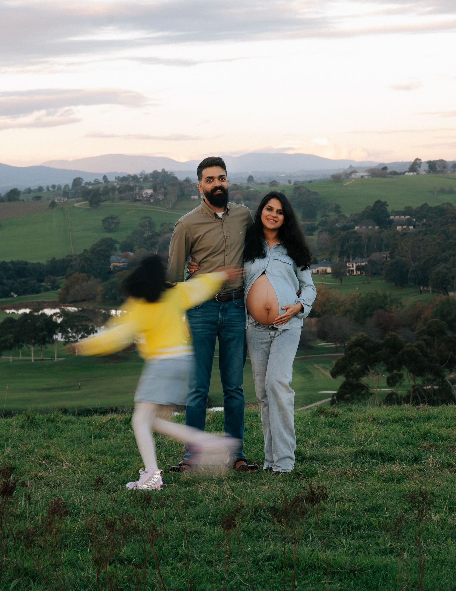 Couple poses with pregnant belly as child runs through green hillside at sunset.