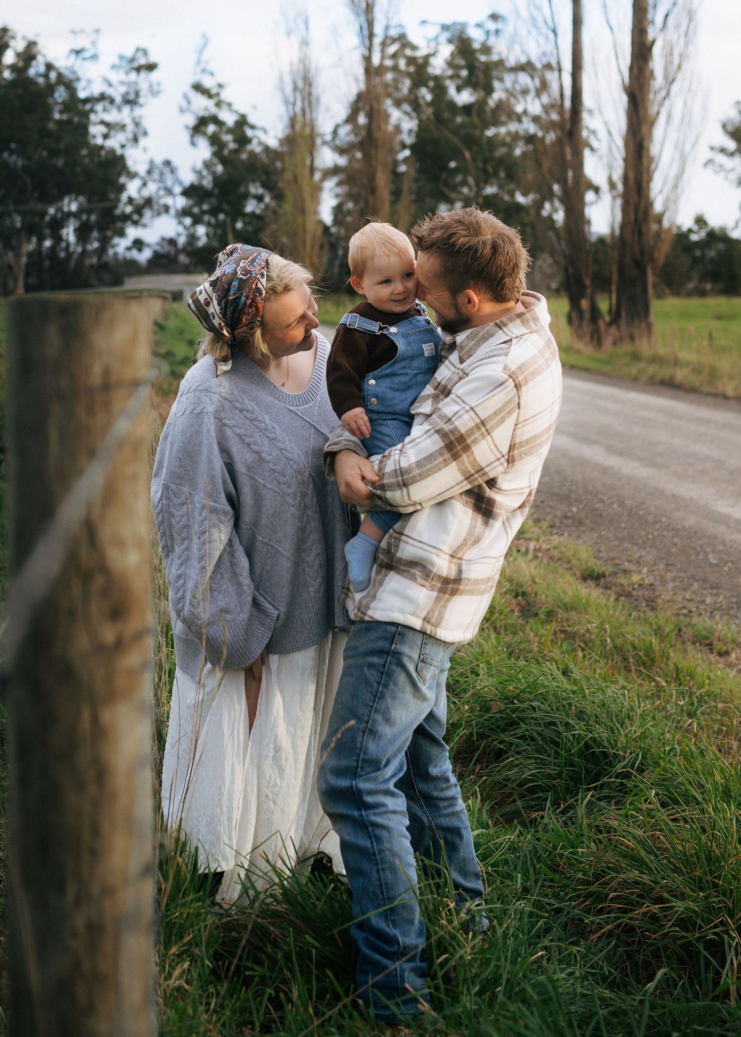 Young family with baby standing by a rustic fence post along a scenic country road.