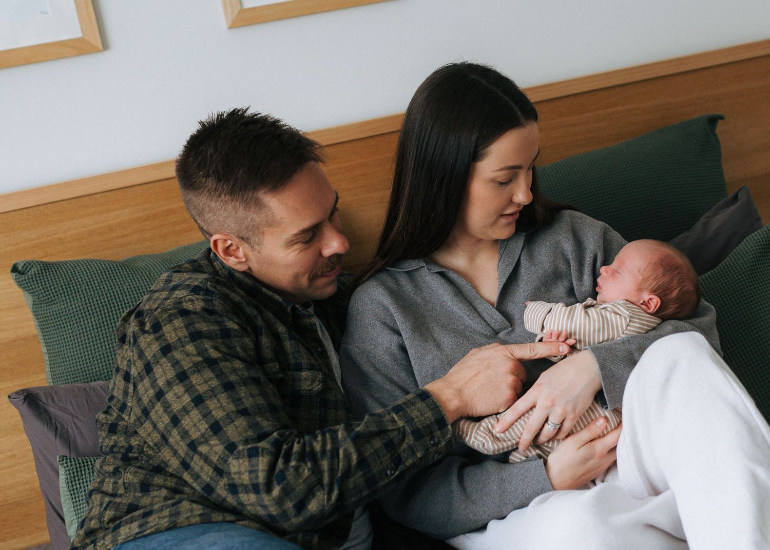 New parents smile lovingly at their sleeping newborn baby while sitting together on a bed.