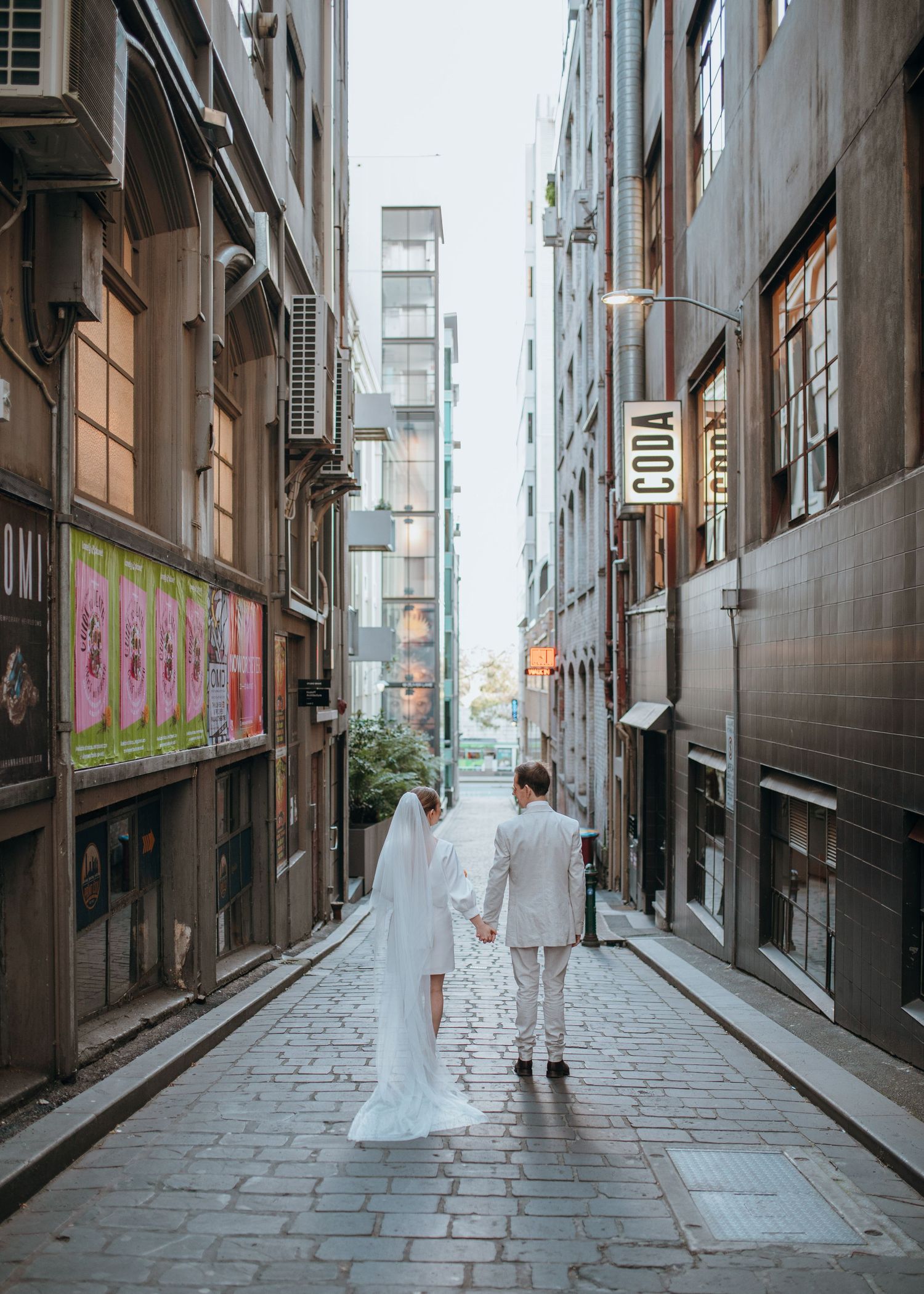 Newlyweds holding hands walking away down narrow cobblestone urban laneway with CODA sign.