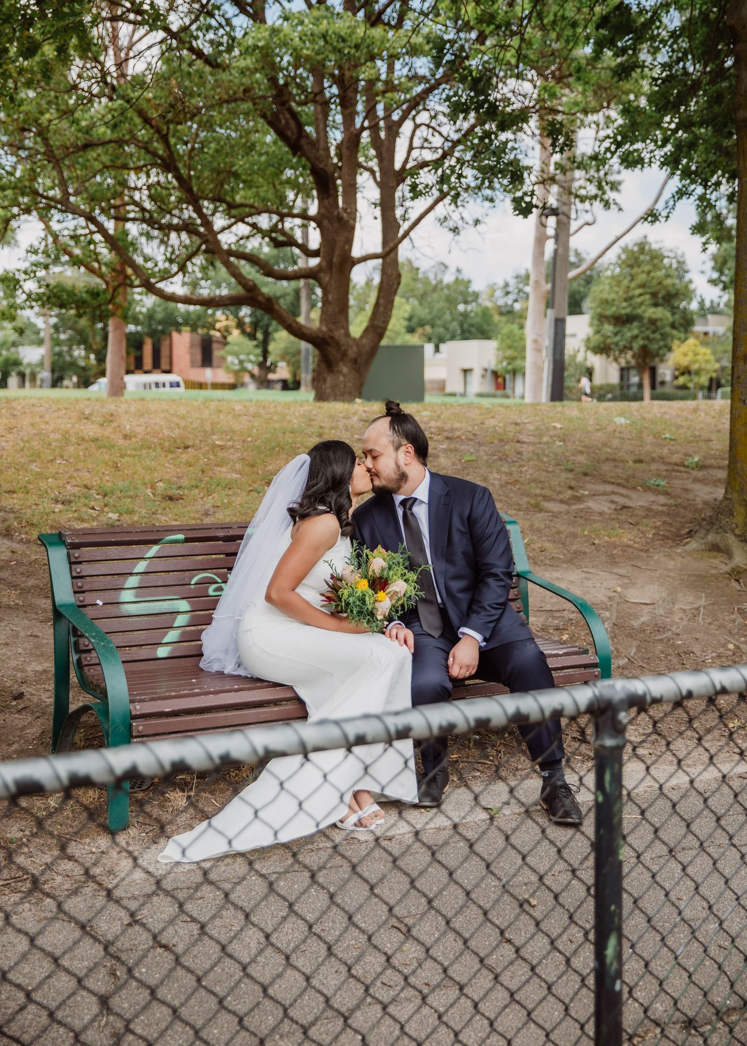Bride and groom kissing on a park bench behind a chain-link fence, bride holding bouquet.