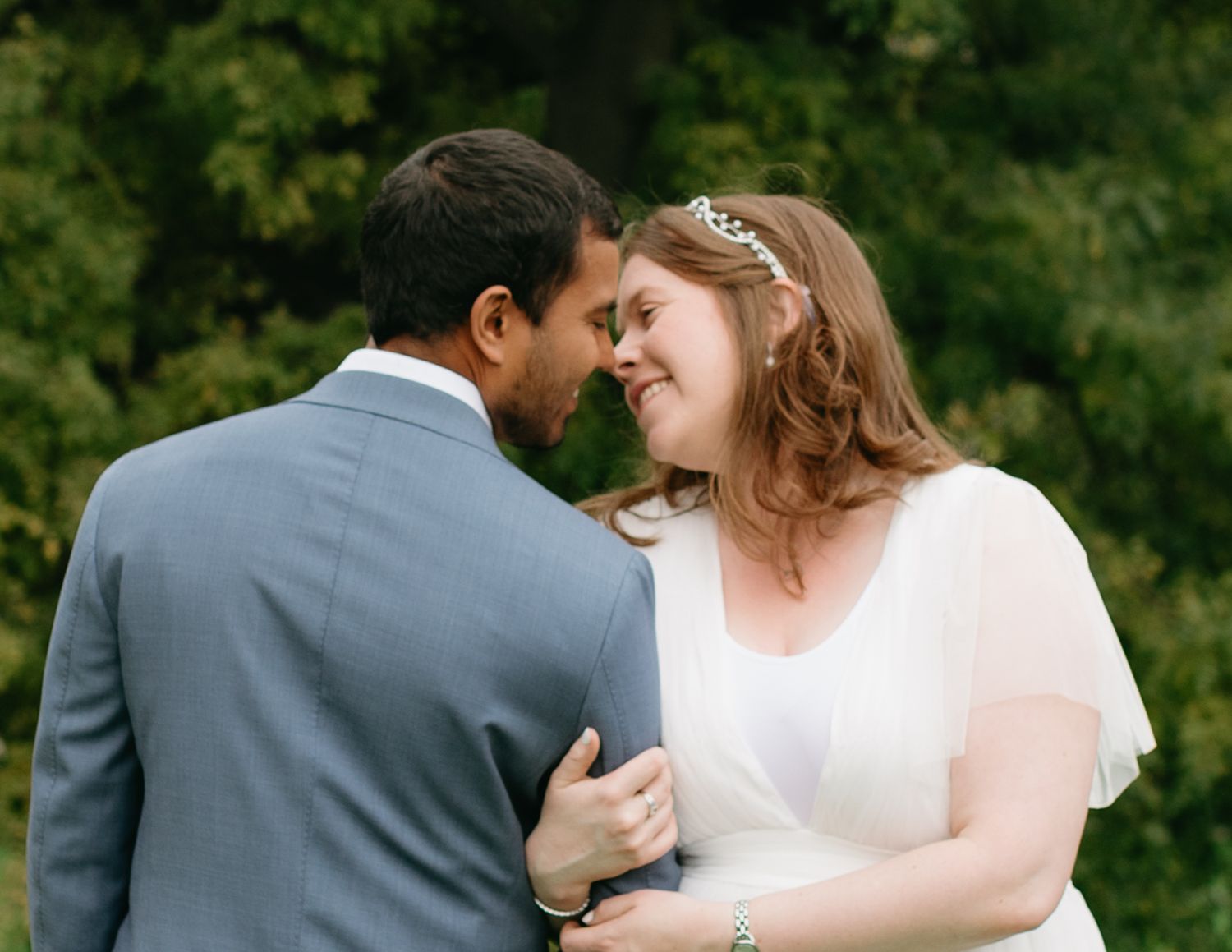 Bride and groom share an intimate moment, smiling at each other outdoors with lush green trees in the background.