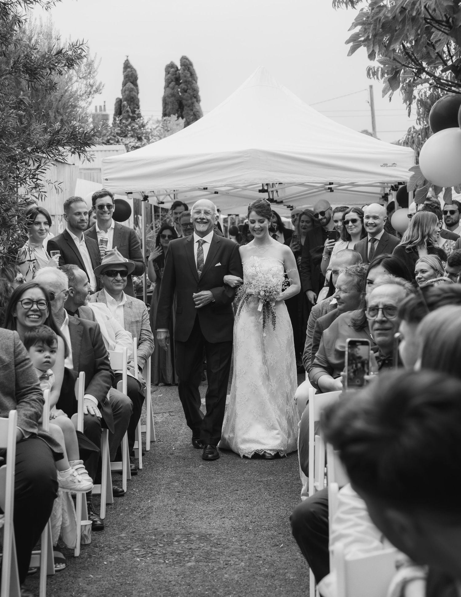 Black and white photo of bride walking down aisle escorted by father at outdoor wedding ceremony.