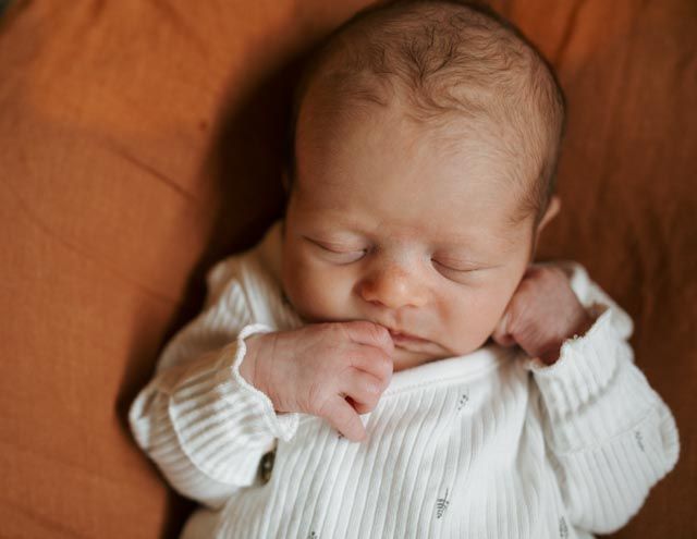 Sleeping newborn baby in white onesie resting on warm terracotta pillow during in-home newborn photography.