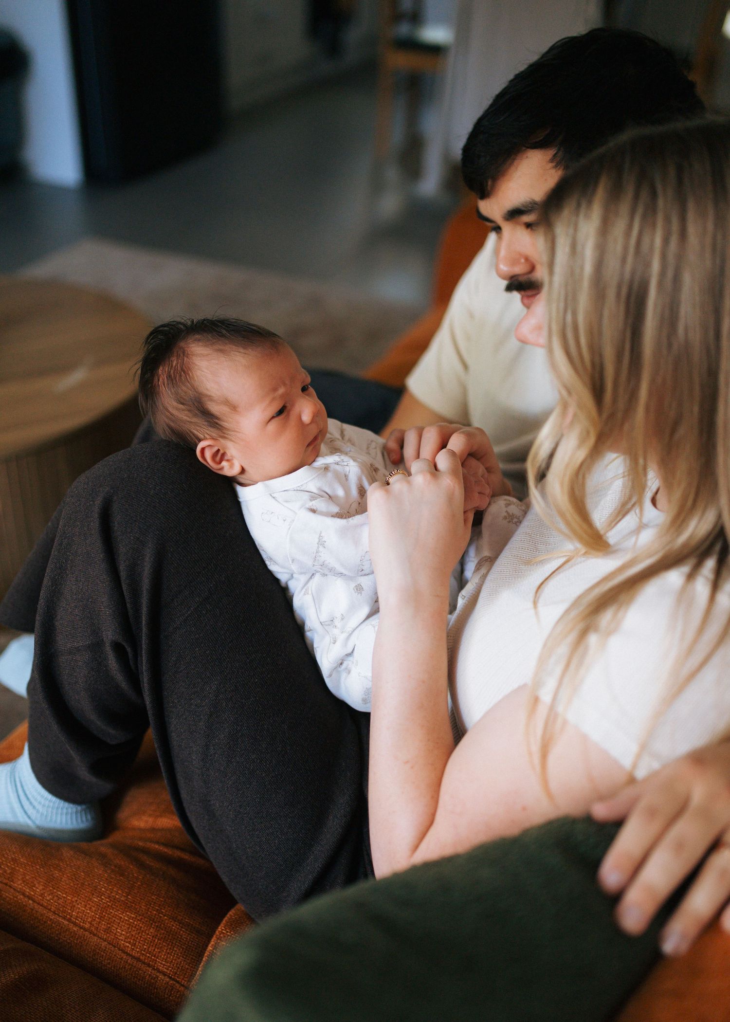 Two parents sit together on an orange couch, gently holding the tiny hand of their newborn baby wrapped in a white outfit.