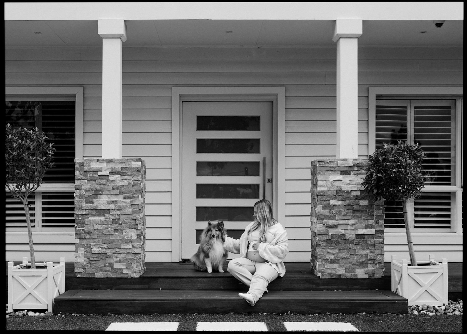 Black and white photo of person sitting on front porch steps with a Collie dog.