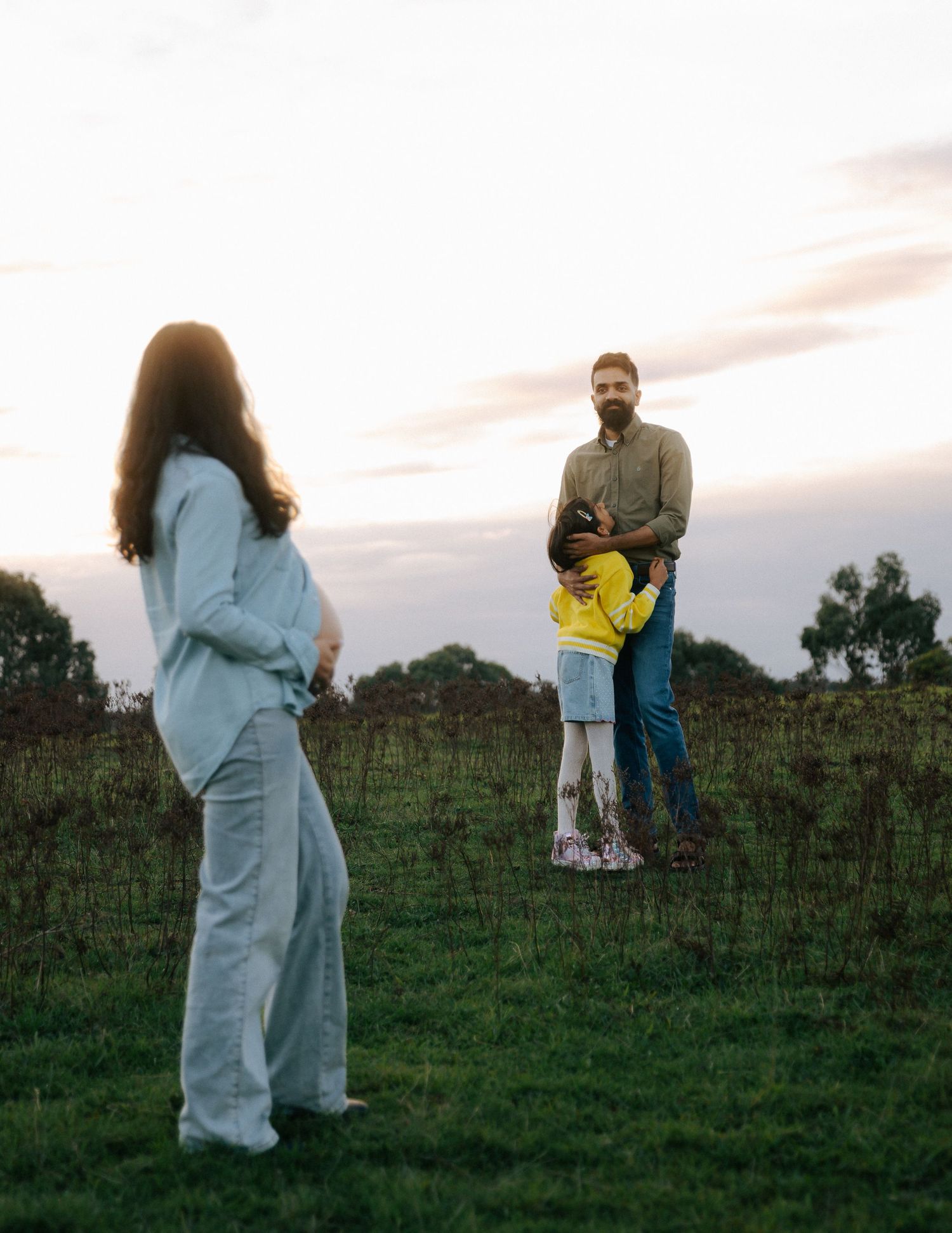 Father holds child in grassy field at sunset during family maternity session.