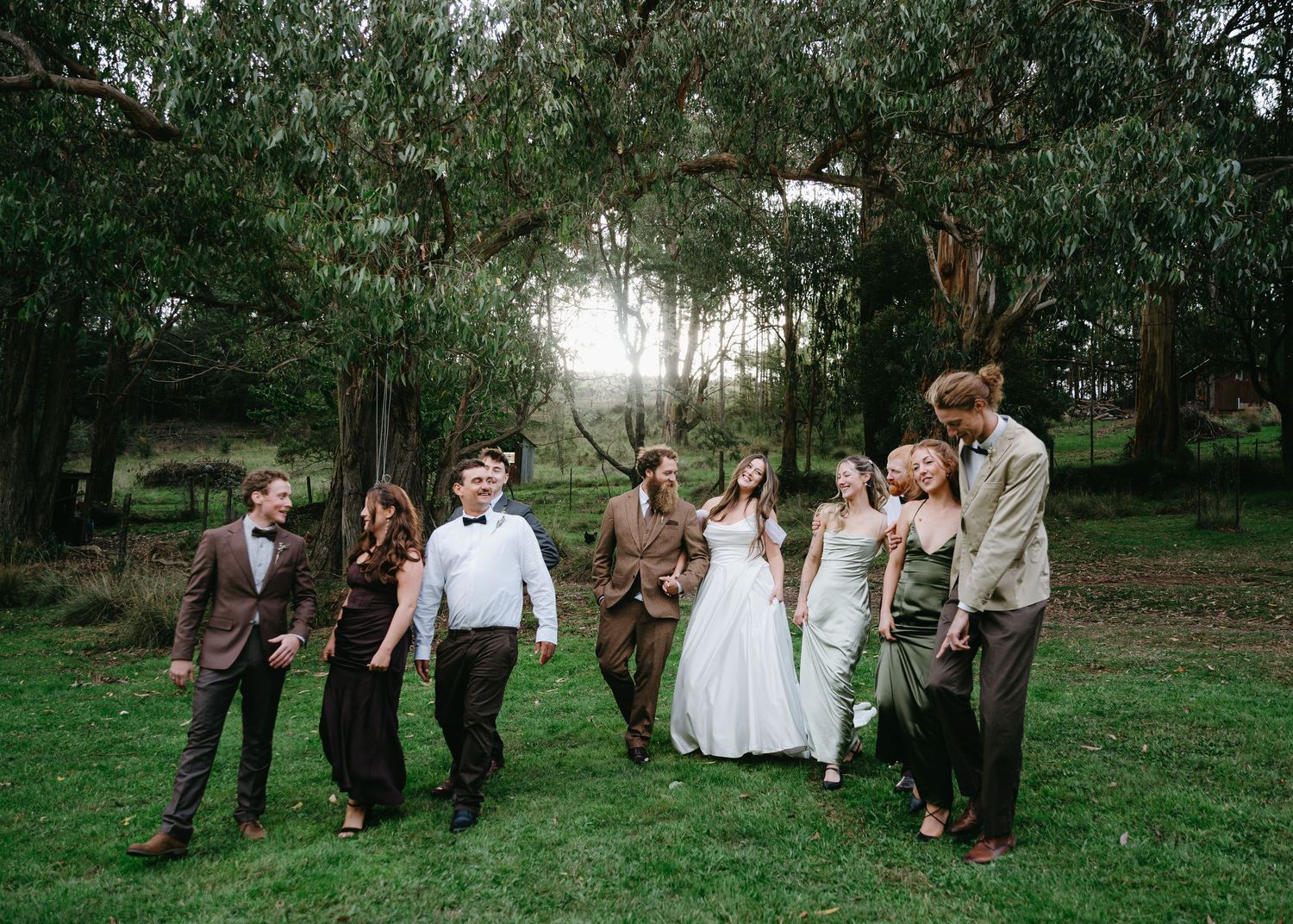 Wedding party walking together through a lush green field with tall eucalyptus trees at golden hour.