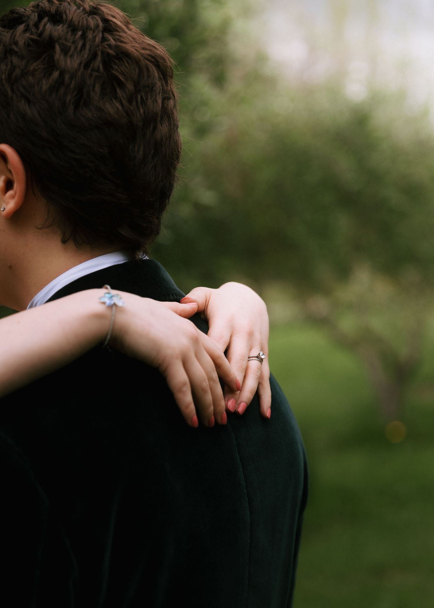 Close-up of a woman's arms wrapped around a man in a dark velvet jacket, a ring on her finger, in a softly blurred green orchard.
