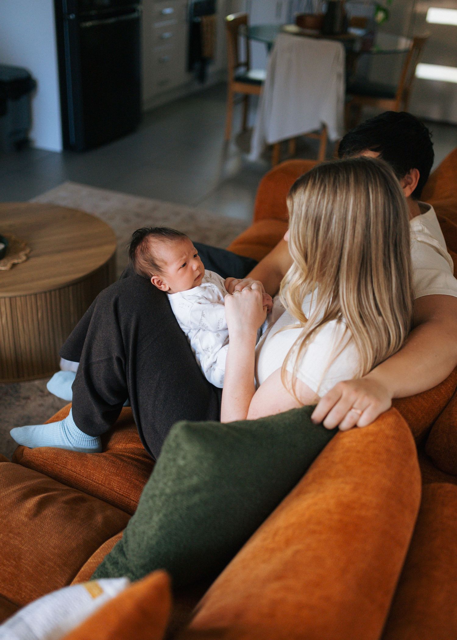 Parents sit together on a cozy orange velvet sofa, gently cradling their newborn baby in a bright, modern living room.