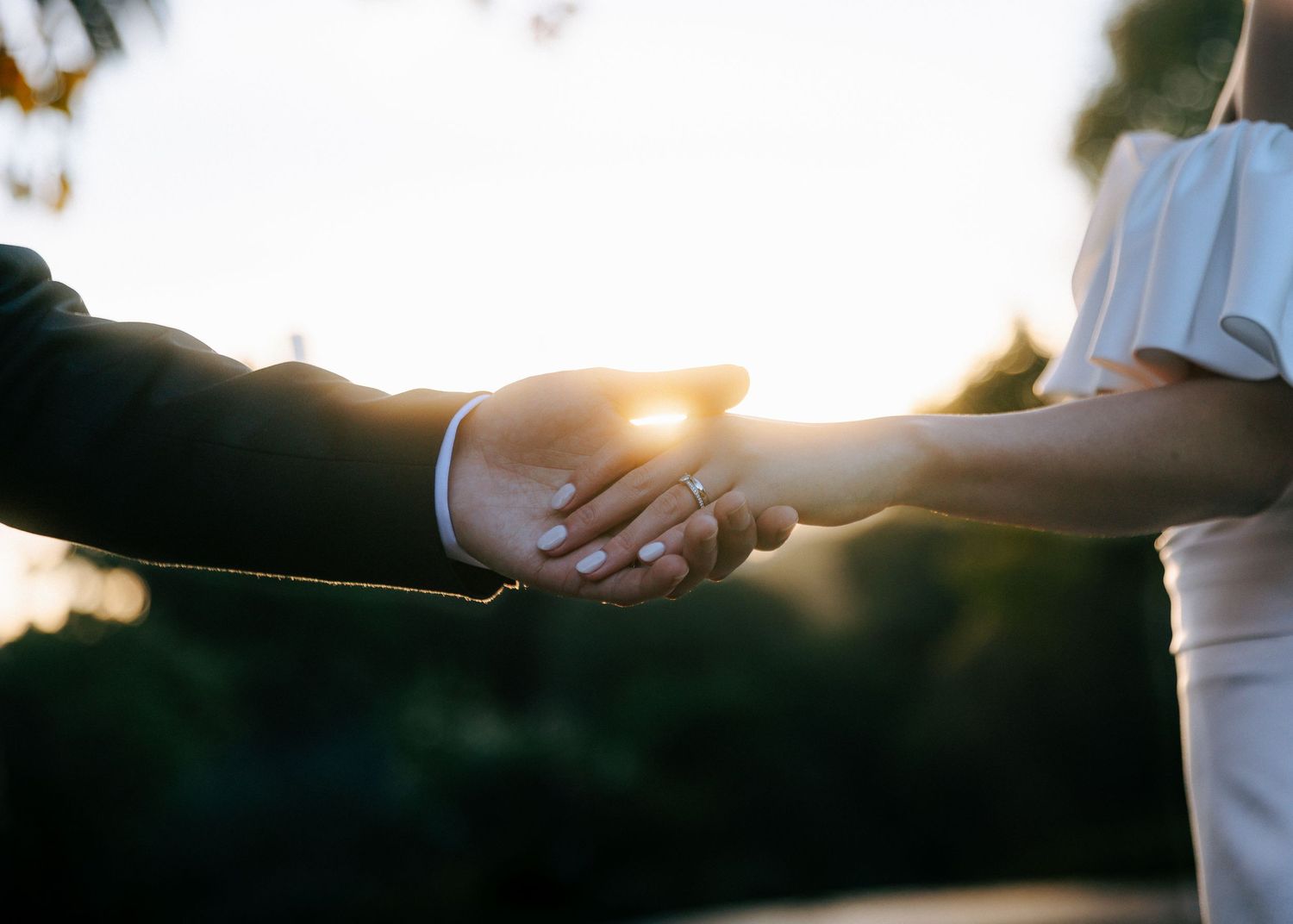 Bride and groom holding hands with wedding rings visible against golden sunset light.