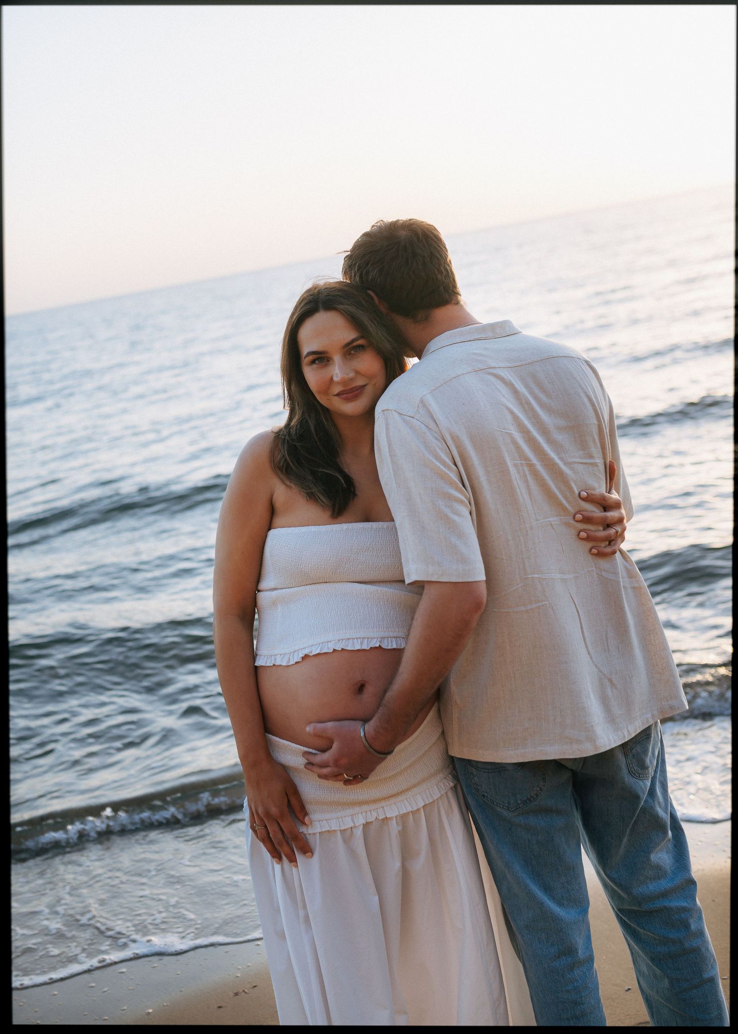Pregnant couple embracing on beach at sunset, woman in white crop top and skirt cradling baby bump by ocean.