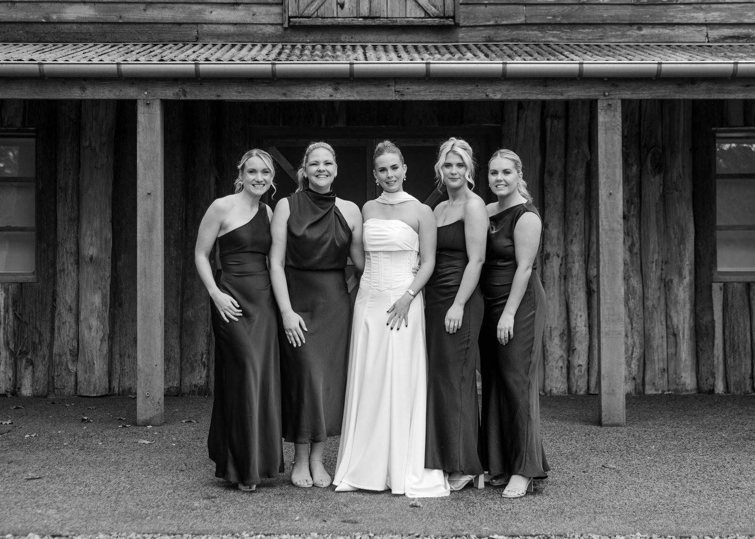 Five women in formal gowns pose together outside a rustic barn, with the bride in white flanked by bridesmaids in dark dresses.