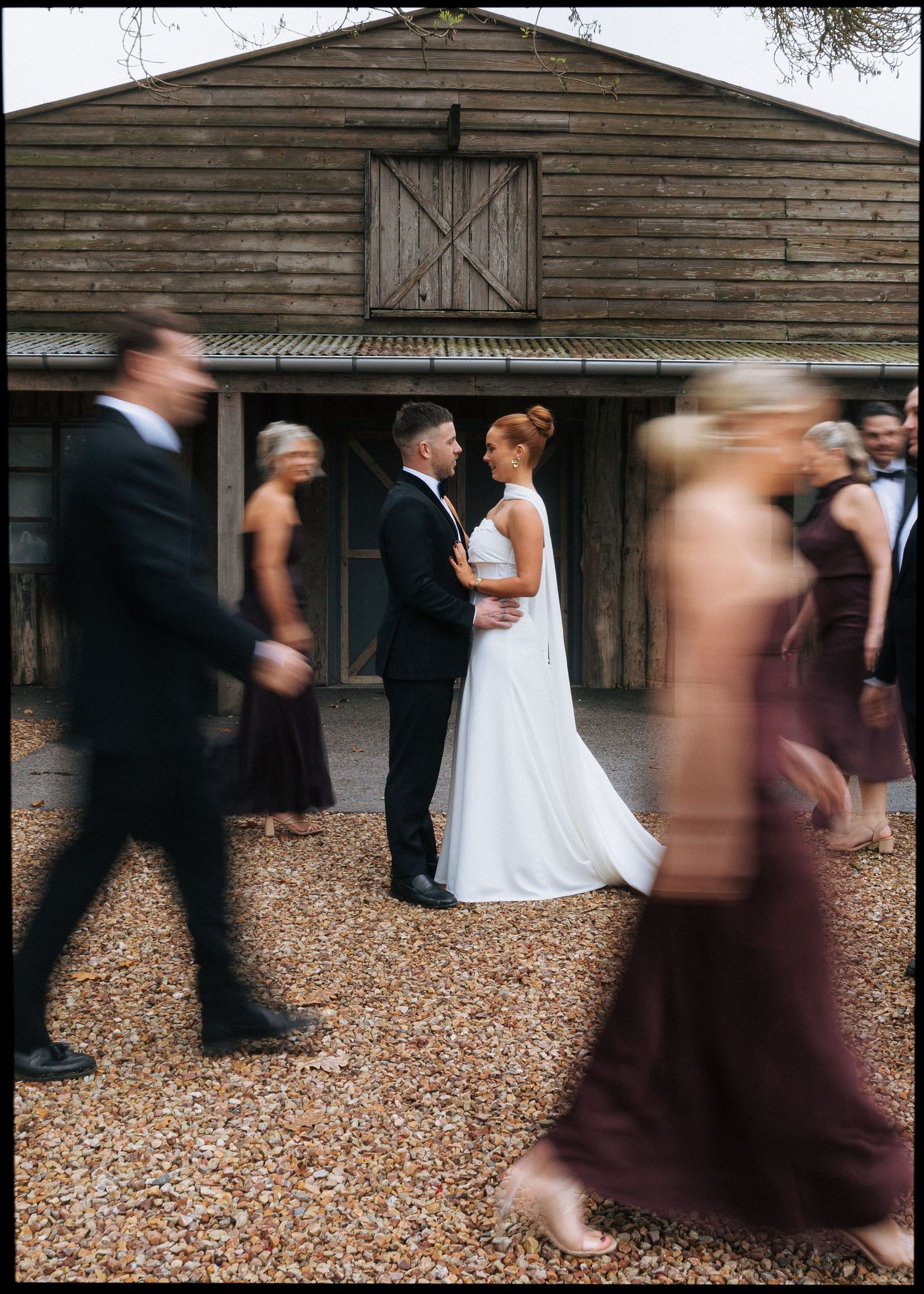 Bride and groom share a romantic moment outside a rustic barn venue as wedding guests walk past in motion blur.