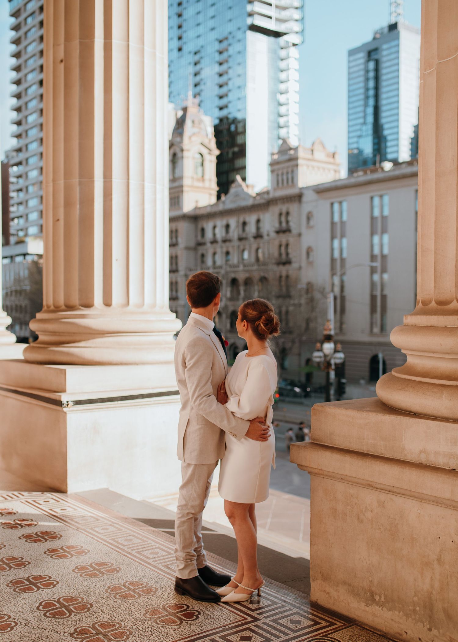 Couple in wedding attire embracing on ornate balcony with city skyline and classical columns backdrop.