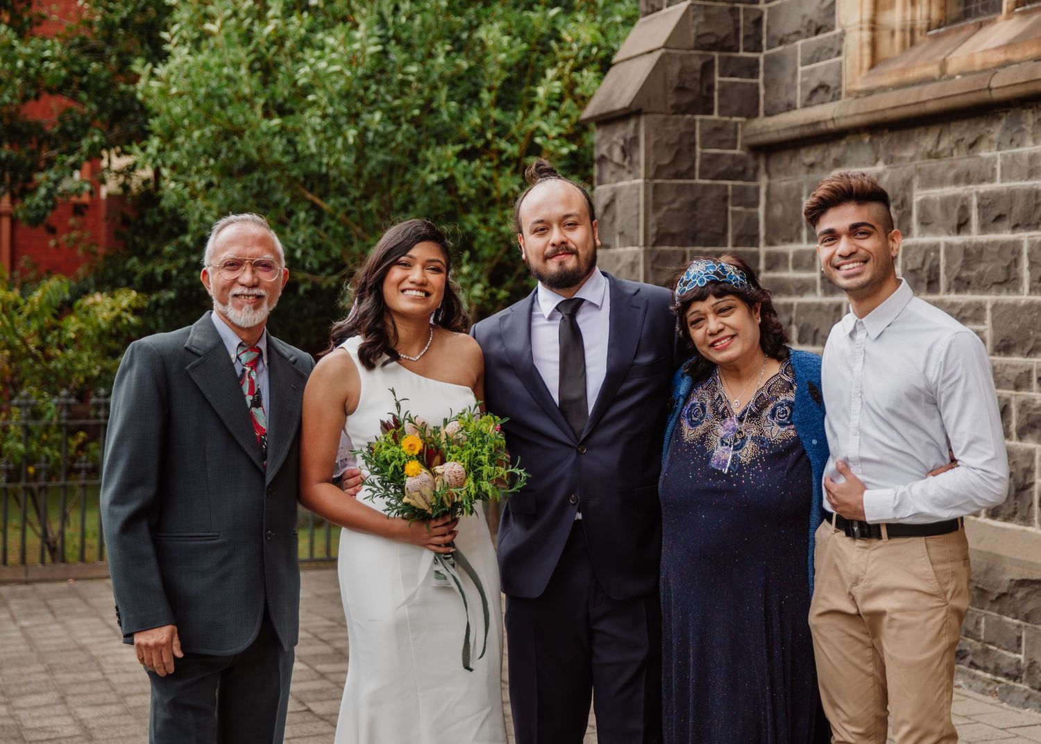 Newlywed couple poses with four family members outside a historic stone church after their wedding ceremony.