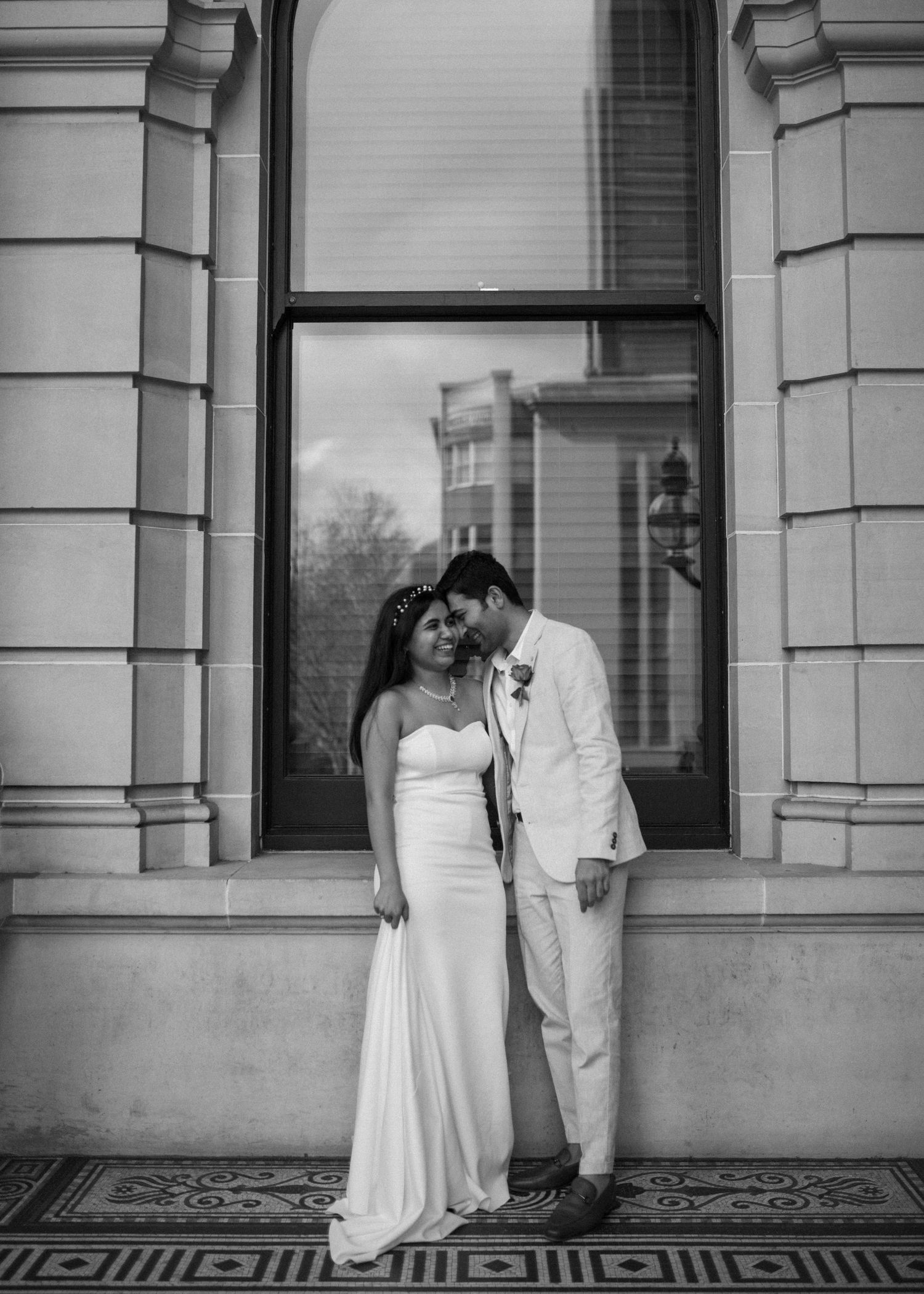 Black and white portrait of bride and groom leaning together by ornate historic building window.