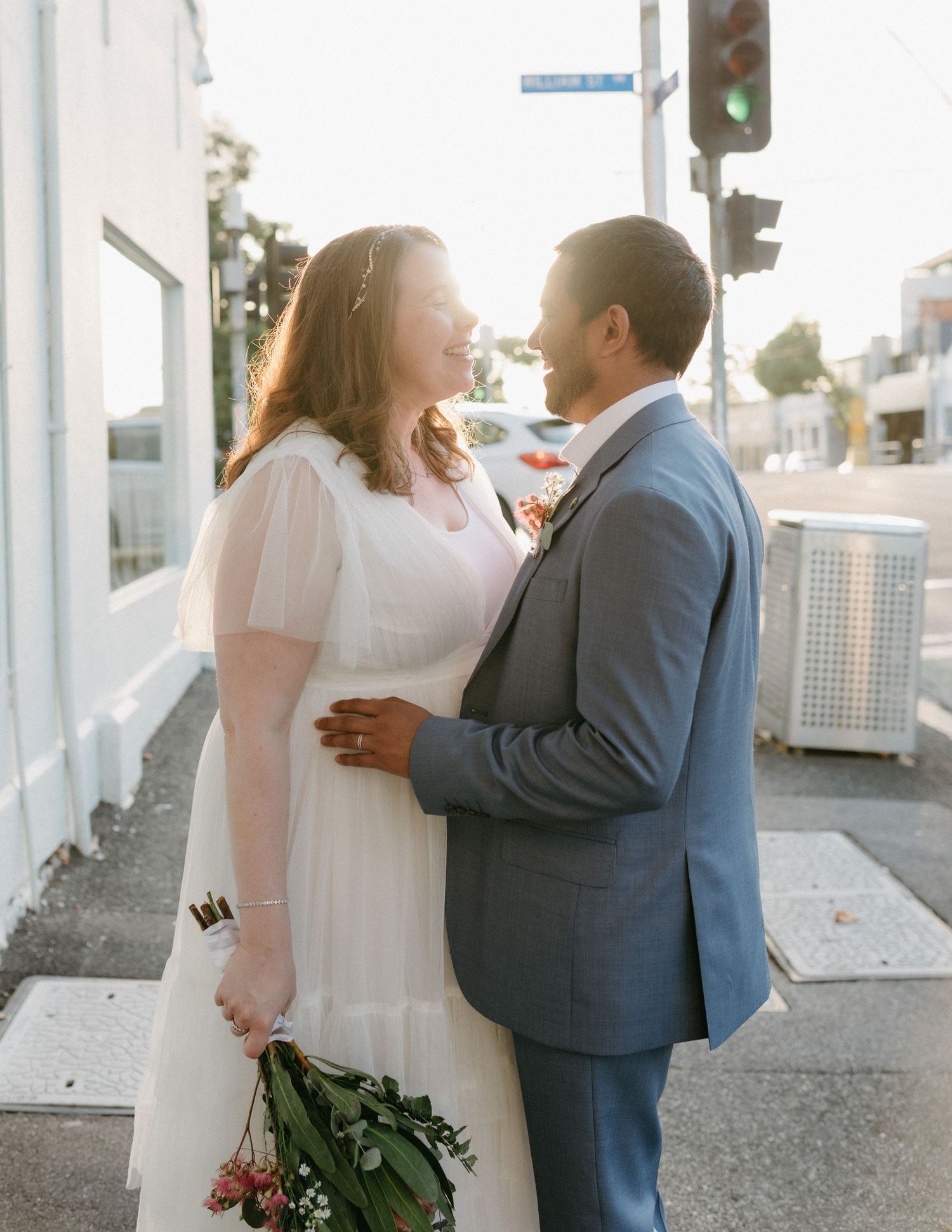 Bride and groom gazing at each other on sunny street corner, bride holding floral bouquet.