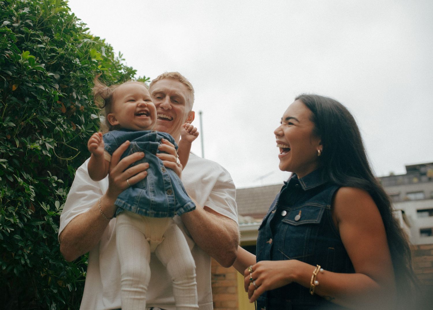 Laughing parents holding joyful baby outdoors in garden with green foliage background.