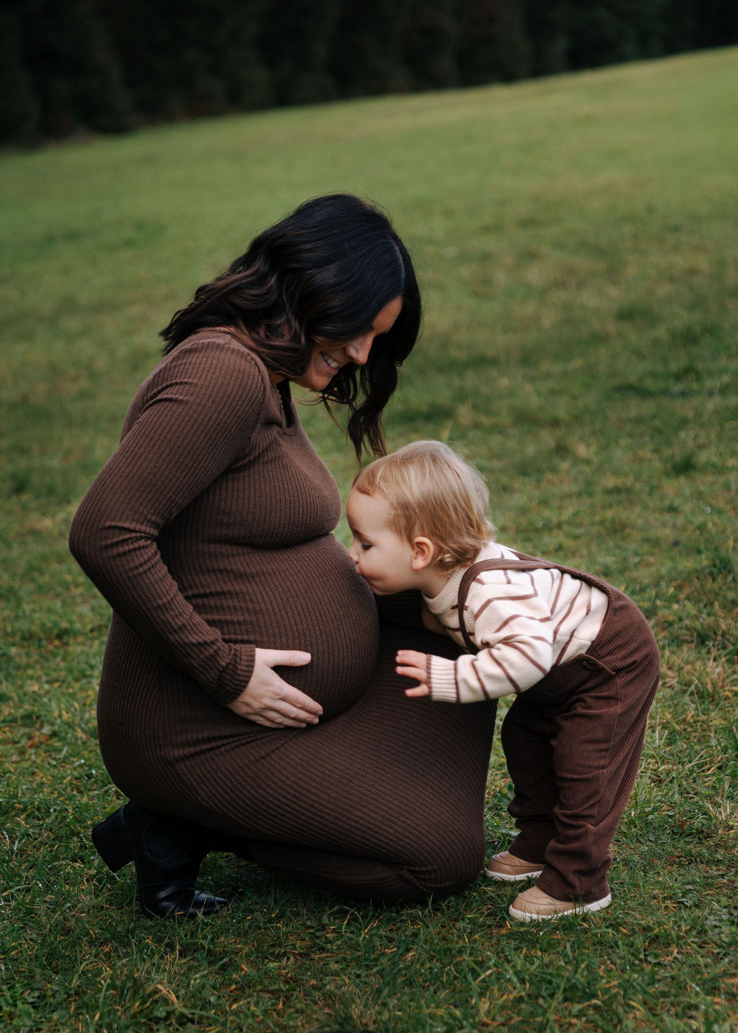 Toddler in striped shirt kisses pregnant mother's baby bump while kneeling on grass.