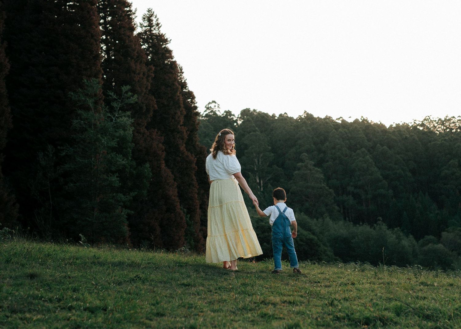 A mother in a yellow skirt holds her young son's hand while walking across a green hillside.