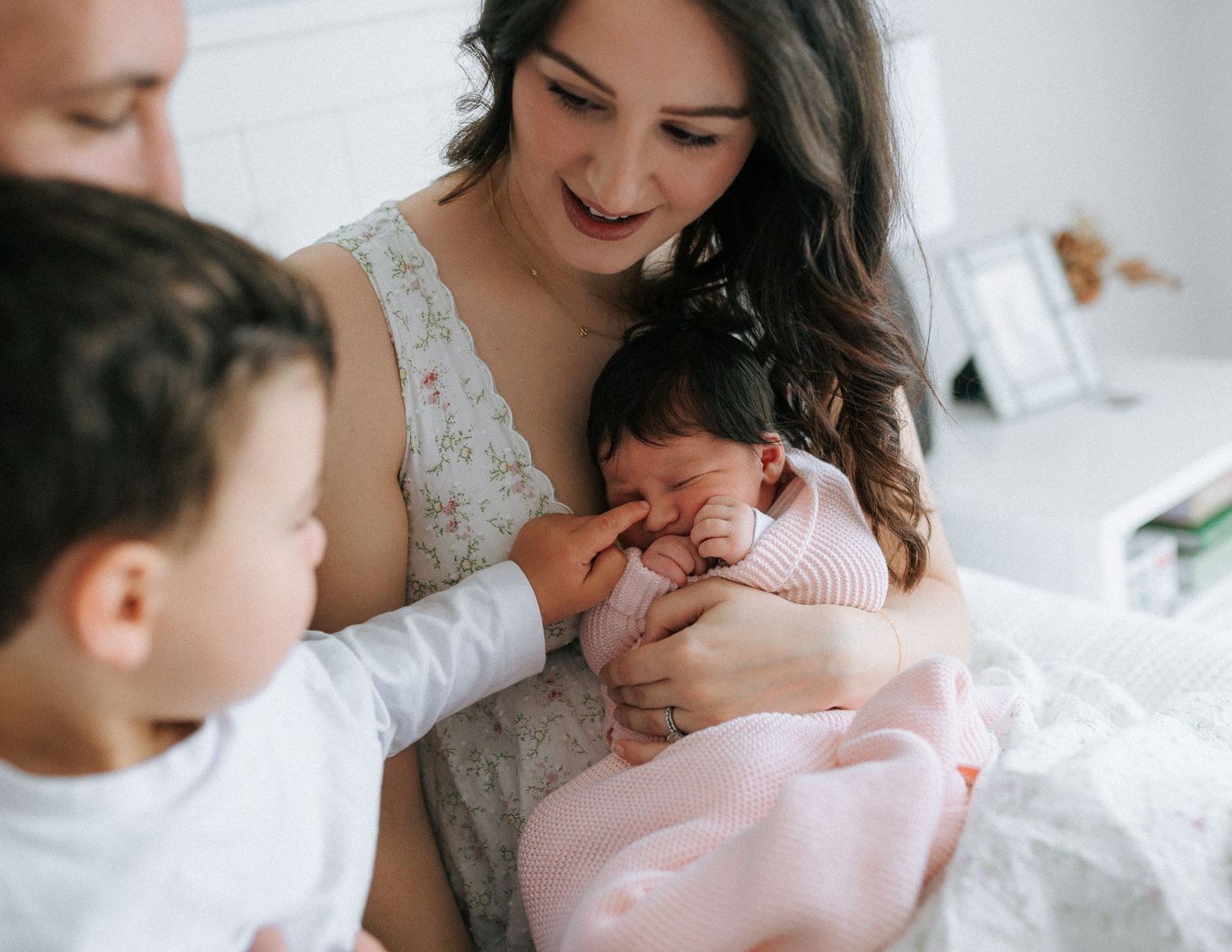 Mother with long dark hair smiling at crying newborn in pink swaddle held by toddler sibling.