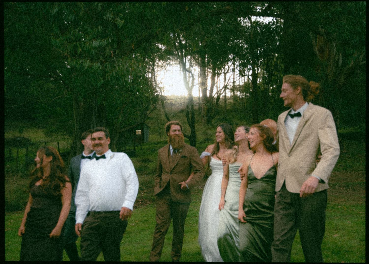 Wedding party walking and laughing together at sunset in a green garden, guests dressed in formal attire.